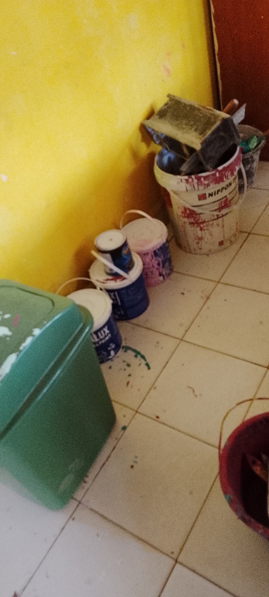 The image depicts a corner of a room with a bright yellow wall and a tiled floor. Several buckets and paint cans are arranged near the wall. In the foreground, a large green plastic container is partially visible on the left. To its right, there are several smaller paint buckets. One is blue with white lettering that appears to say "LUX". Another is pink and patterned. A larger white bucket with red markings and the brand name "NIPPON" is further back, containing some tools like trowels. Splatters of paint, in red and green, are visible on the floor. The overall scene suggests a space where painting or renovation has recently taken place or is in progress. The image was taken in Jepara, Indonesia.