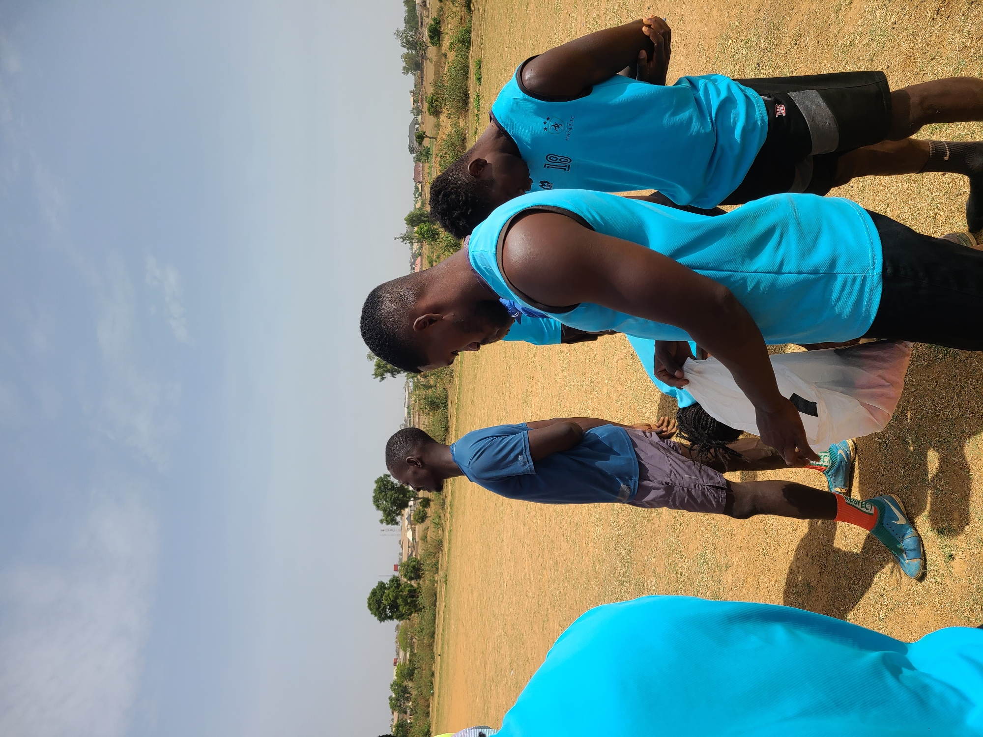 A group of young men, likely athletes, are gathered outdoors on a dry, earthy field under a bright, clear blue sky with sparse clouds. The scene appears to be set in Barikin Jirga Summa, Nigeria.
The majority of the men are wearing light blue sleeveless athletic shirts, some featuring "AVANJEL FC" and a logo resembling three stars on the back, with one clearly displaying the number "18". They are also wearing dark shorts. One man is dressed in a darker blue t-shirt and grey shorts. Many have their heads bowed, suggesting a moment of solemnity, prayer, or focused attention, possibly before or after a sports event. One individual is reaching into a white plastic bag. Their postures indicate a moment of rest or instruction.
Footwear includes athletic shoes, some appearing to be football (soccer) cleats/turf shoes, and athletic socks. One pair of orange socks prominently displays the word "RESPECT" in white lettering.
In the background, the field extends to a distant treeline, beyond which sparse buildings and structures are visible, hinting at a rural or semi-urban environment. The time of day is bright, likely midday or afternoon, as indicated by the clear lighting and visible shadows. The overall atmosphere suggests a pause in an athletic activity on a sunny day.