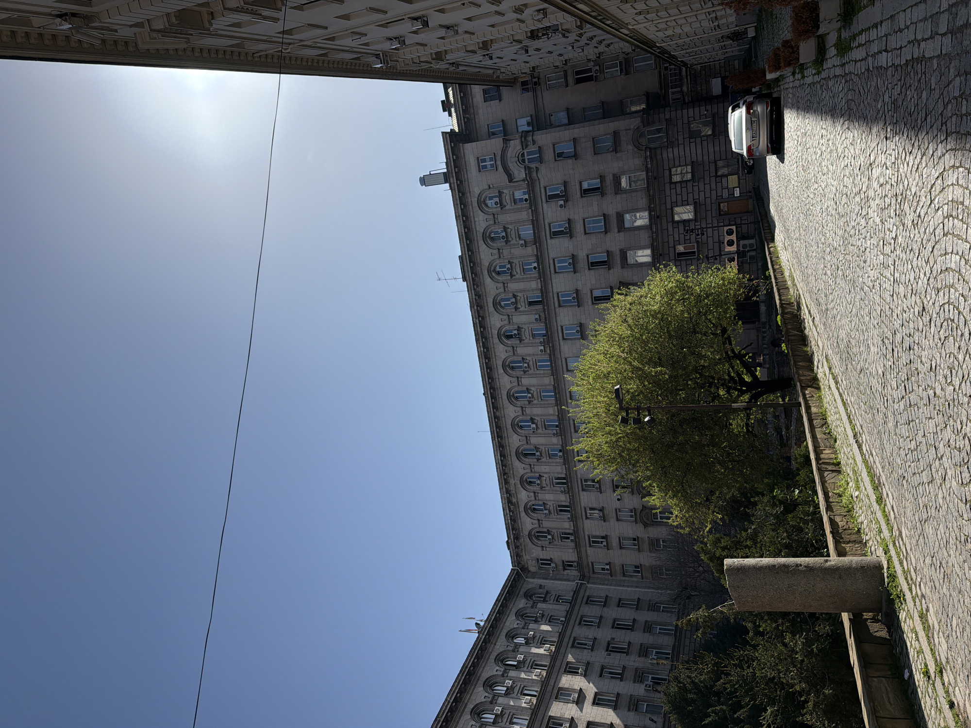 This image captures a sunlit urban street in Sofia, Bulgaria, characterized by historical architecture and a clear sky. A prominent, elongated, grey building, several stories high, dominates the right side of the frame. It features numerous windows, some with arched frames, and is adorned with architectural details like cornices and multiple air conditioning units on its facade. The ornate cornice of another building is partially visible on the far left.
A cobblestone street, partly illuminated by sunlight and partly in shadow, curves from the foreground towards the upper right. A silver car with the Bulgarian license plate "CA 1718 MP" is parked on this street in the upper right quadrant. A large, verdant tree with thick foliage stands in the mid-ground, partially obscuring the building behind it, with additional greenery at ground level. A sturdy, grey stone pillar or bench is visible next to the street in the lower right.
The sky is bright and clear, indicating a sunny daytime. A sun flare is visible in the upper left portion of the sky, and a thin power line diagonally traverses the upper part of the scene. The overall impression is of a quiet, old-world European city street on a bright day.