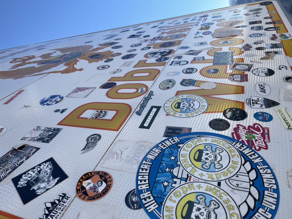 The image displays a large, reflective white surface, likely a roadside sign or monument, situated outdoors under a clear blue sky. The surface is extensively covered with numerous stickers of varying sizes, shapes, and designs. Prominently featured in large, golden-yellow, outlined letters are the words "Добродошли" (Dobrodošli, meaning "Welcome" in Cyrillic) and partially visible "Welcome" in Latin script below. A large golden double-headed eagle with a shield on its chest, consistent with national symbols of the region, is affixed to the upper right portion of the sign.
The stickers present a diverse collection, primarily associated with motorcycle clubs, rallies, and automotive interests. Notable examples include "BIG RALLY CROATIA 06," "MOTO VINTAGE SUD," "CHAUSSER ATV & MOTO," "BOSS TRUPPE," "Street Gang Rodrigo," and "Super G Team." Two prominent circular blue stickers in the lower-left feature a skull wearing a traditional cap, crossed bones, and Cyrillic text "ЦРНА ГОРА" (Crna Gora, Montenegro), along with German text "HIER-REGIERT-NUR-EINER" ("Only one rules here"). Many smaller, less legible stickers also contribute to the dense coverage. The background features a clear blue sky in the upper right quadrant, with a blurred, distant landscape or mountains visible along the far right horizon. This scene is located in Klobuk, Bosnia and Herzegovina.