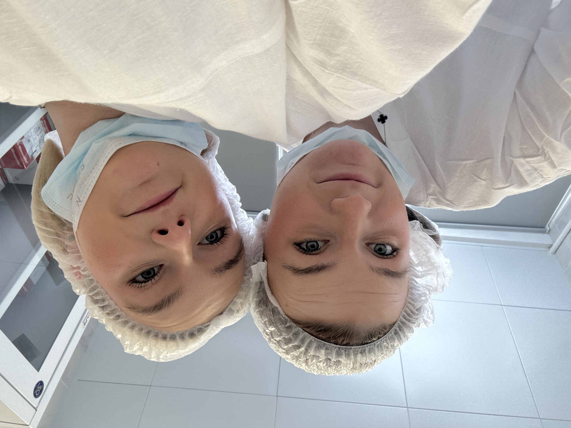 Two individuals wearing medical masks and hairnets stand in close proximity. Both are clad in white medical gowns, possibly surgical scrubs. They are positioned indoors, where the background reveals a tiled wall and a white cabinet. The location is Saint Petersburg, Russia. The individuals seem to be looking down at the viewer.