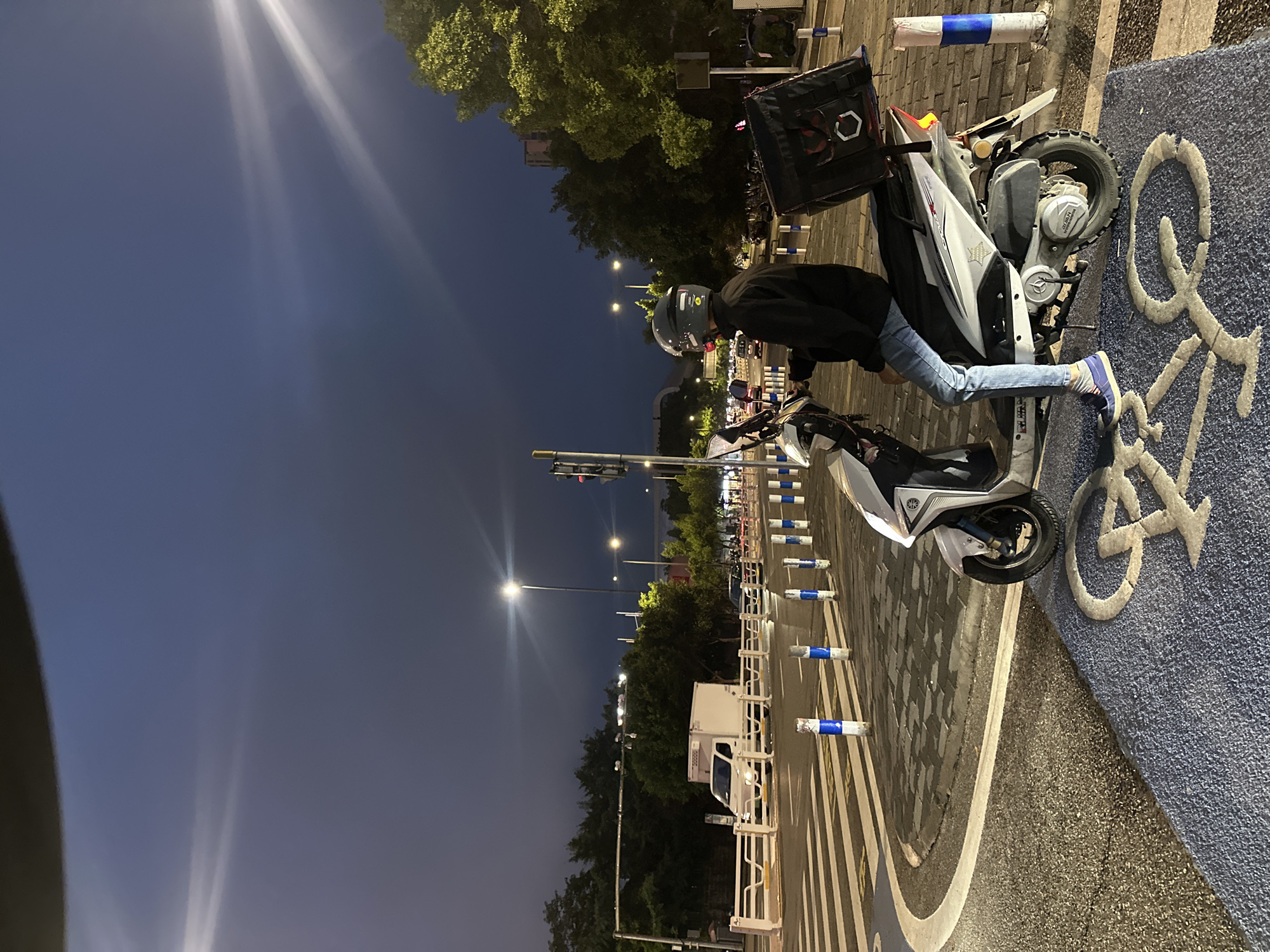 A person wearing a black jacket, blue jeans, a grey helmet, and grey socks with blue shoes is sitting on a white scooter. The scooter has a black storage box on the back. The person is looking to the right, away from the camera. The scooter is parked on a blue mat with a white bicycle symbol on it. The scene appears to be a street intersection at night. There are street lights and traffic lights visible in the background, along with trees and buildings. The ground is wet, suggesting it might have recently rained. The location is specified as the city of Chongqing High-tech Zone, China.