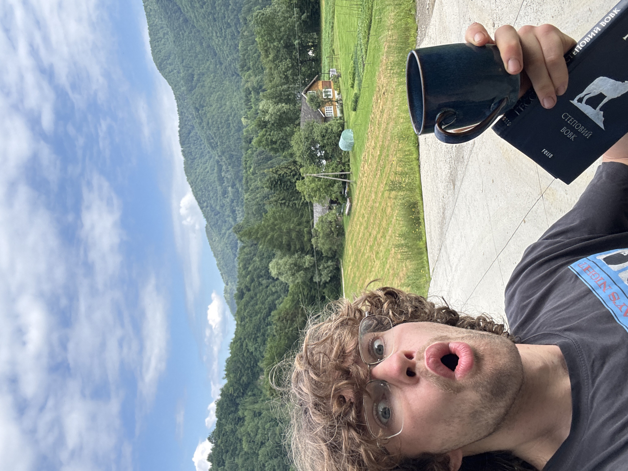 A young man with curly brown hair and glasses is taking a selfie outdoors. He is wearing a dark t-shirt. His mouth is open in a surprised expression. He is holding a dark blue mug in his right hand and a dark book in his left hand. In the background, there are green hills covered in trees, a grassy field, a house with a red roof, and a greenhouse. The sky is blue with scattered white clouds. The location is described as Unknown, Unknown.