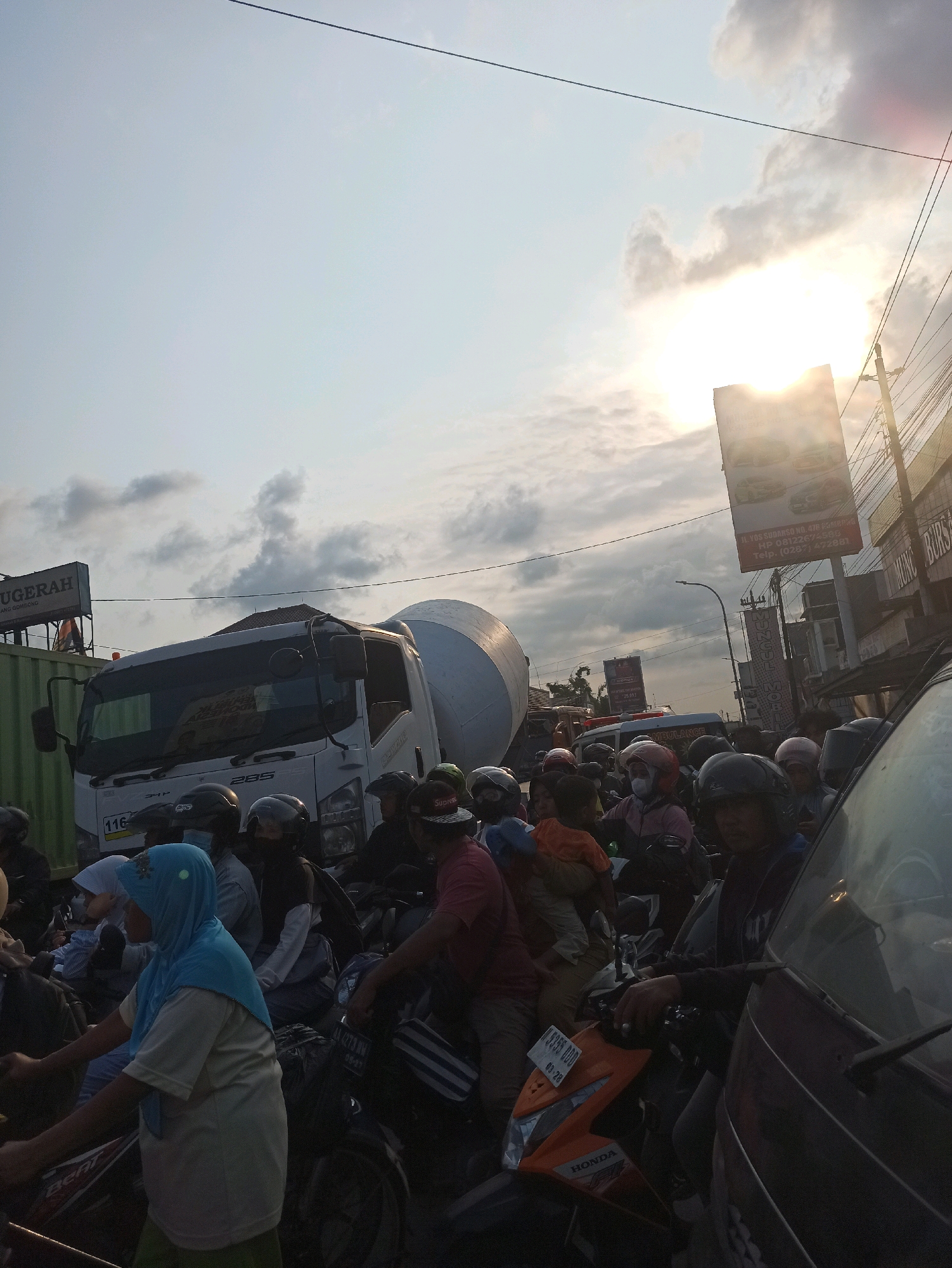 The image depicts a traffic jam in Gombong, Indonesia, with numerous motorcycles and a cement mixer truck on a road. The sky is overcast with the sun peeking through the clouds, suggesting it is late afternoon or early evening. Several people are visible, mostly on motorcycles, some wearing helmets and others with head coverings. A large green container is on the left side of the frame, and various signs are posted on buildings in the background. The scene is crowded and appears to be a typical urban environment with a mix of vehicles and pedestrians.