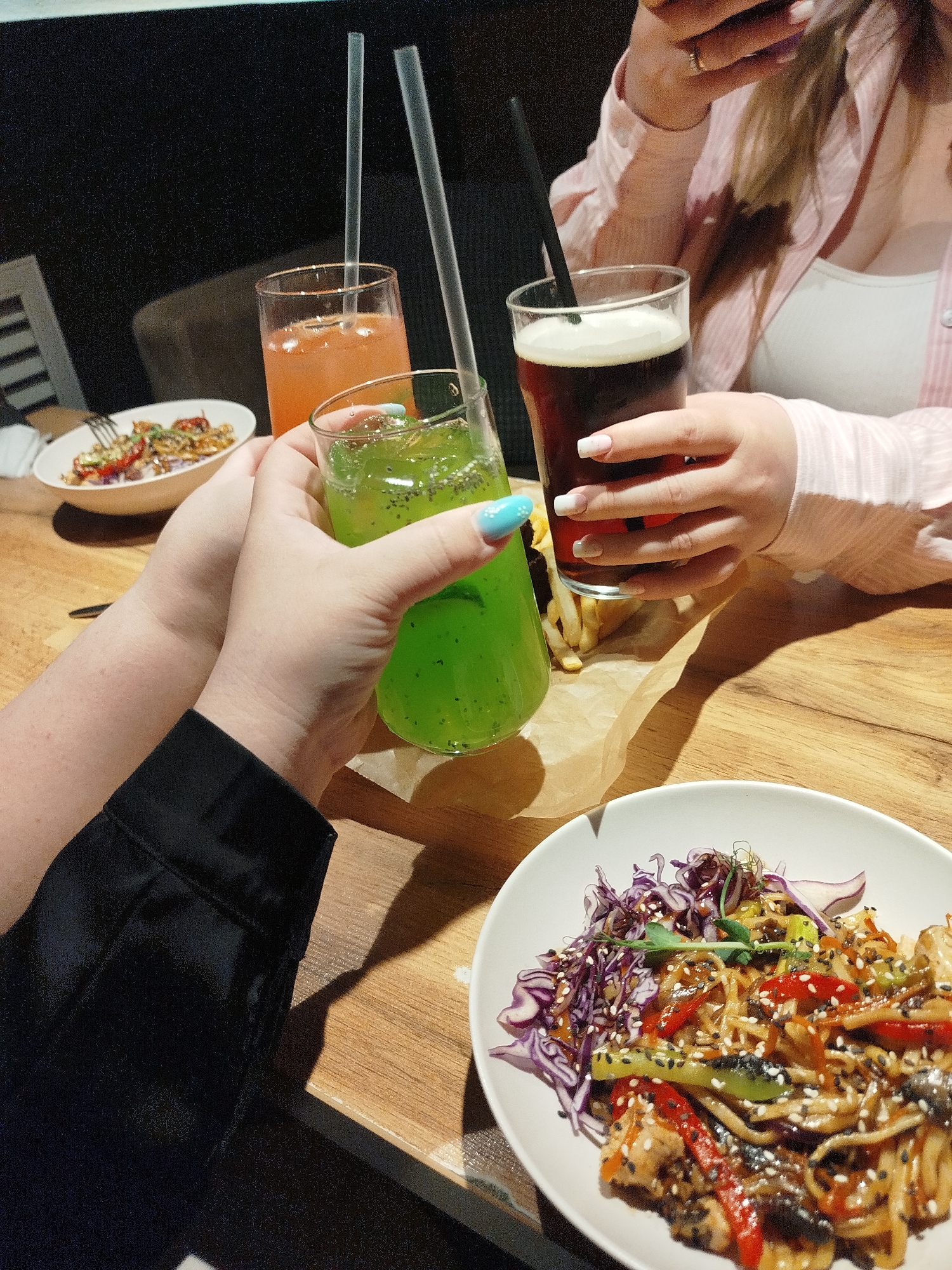 Two people are at a wooden table in what appears to be a restaurant or cafe. They are toasting drinks - one green, one orange, and one dark red - with straws visible. One person is wearing a black garment, and their nails are painted with a pattern including a light blue color. The other person is wearing a light pink button-down shirt over a white top. On the table are two plates of food, one with noodles, vegetables, and sesame seeds, and the other with a mixed salad-like dish. A piece of paper is visible under the drinks, possibly holding french fries. The scene is indoors and illuminated with artificial light. The location is Chaikovsky, Russia.
