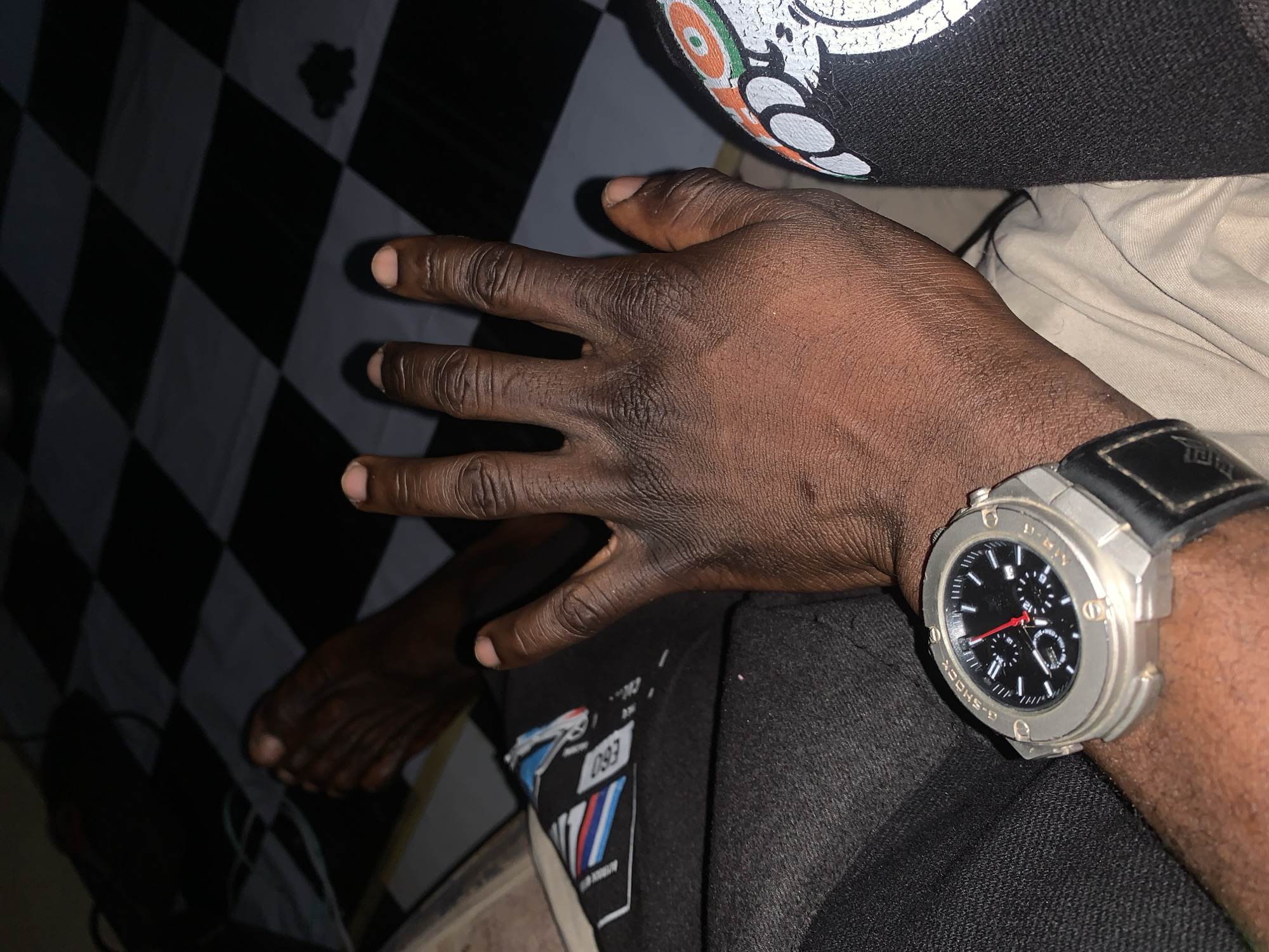 The image shows a close-up view of a person's hand, foot, and a watch. The hand is extended with fingers spread, resting on a light-colored surface. The foot is visible in the background, appearing to be bare. A silver-colored watch with a black strap is worn on the wrist. The background consists of a black and white checkered pattern, possibly a floor or a mat. The lighting suggests an indoor setting at night. The image's context in New Stadium, Nigeria, is not discernible from the visual information provided.