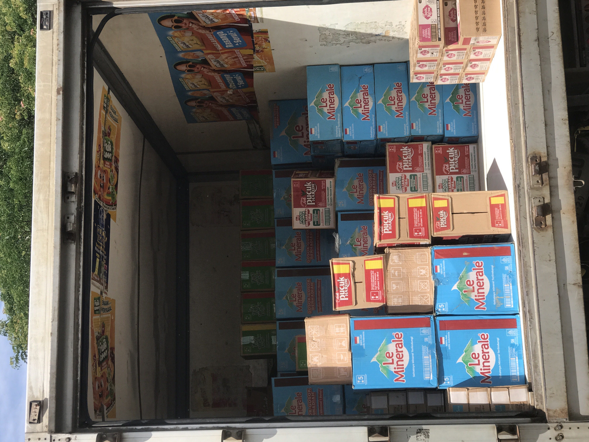 The inside of a truck is filled with boxes of drinks. The brand "Le Minerale" is prominent on blue boxes stacked throughout the truck. There are also brown boxes labeled "Teh Pucuk Harum." The inside walls of the truck have posters advertising various food and drink products, including "Frutta Gummy" and "Durian Gelato." The overall environment appears to be a delivery or storage space for beverages, with the goods packed tightly inside the truck. Given the brands and the OCR, it is likely that the truck is in the city of Miruektaman, Indonesia.