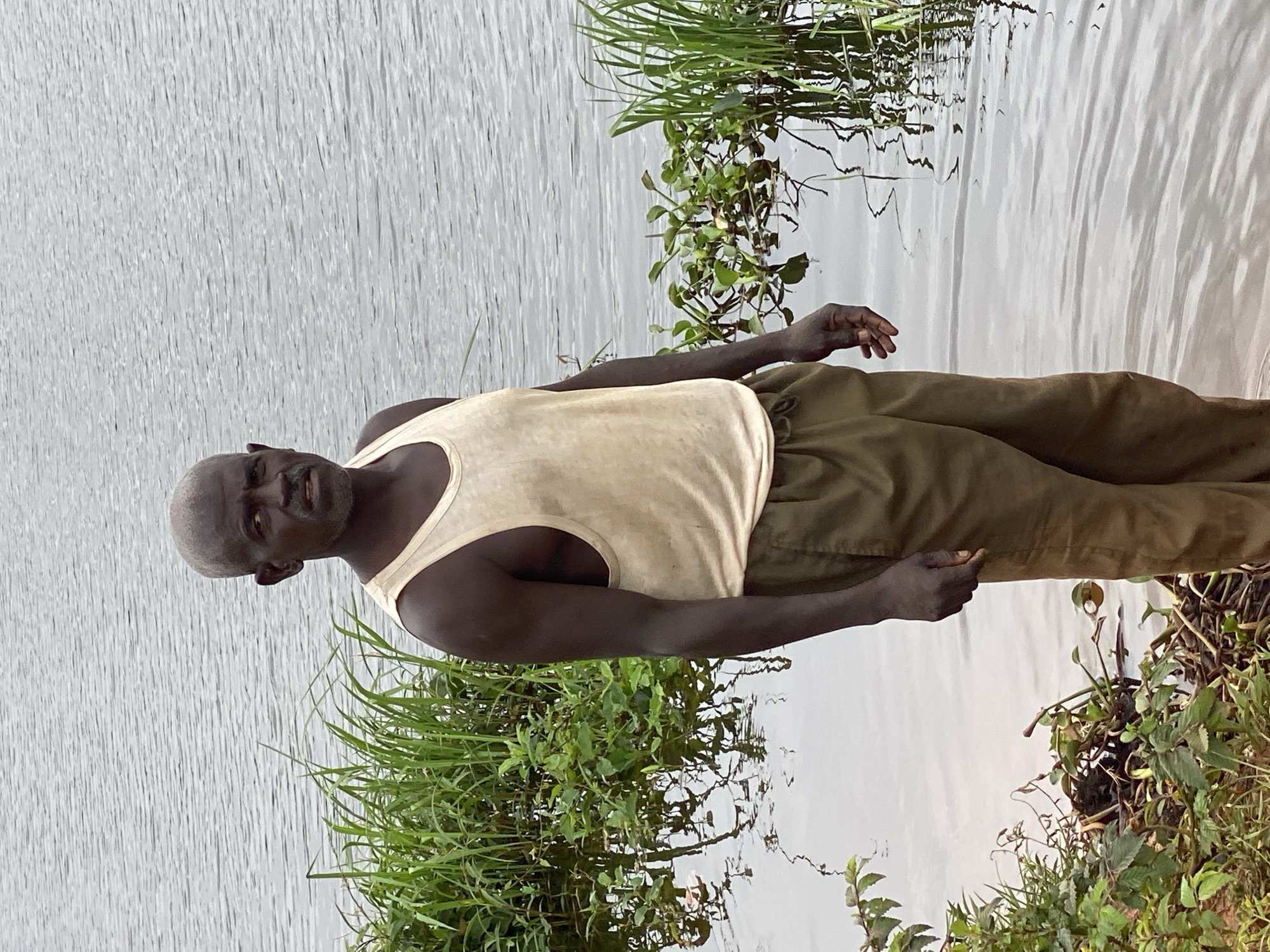 A medium shot captures a dark-skinned man standing waist-deep in murky water near the shore of a body of water. He has a shaved head and is wearing a tan tank top and olive green pants. The man is looking directly at the camera with a serious expression. The background consists of calm water with ripples, and on either side of the man, there are patches of tall green grass and aquatic plants. The scene is likely in Kawo, Nigeria.