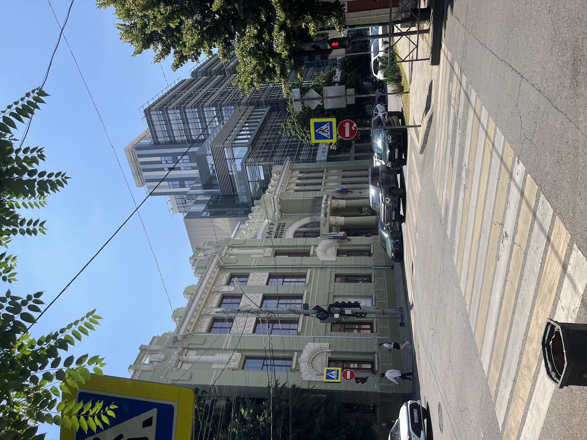 The image depicts a street scene in Krasnodar, Russia, featuring a prominent light green, ornate building with classical architectural elements on the left, likely a theater given the visible signage that reads "ТЕАТР ШАТНАЯ ЕЩЕСТВА" (Theater Shatnaya Echestva). Adjacent to and behind this older structure is a modern, multi-story glass and steel building, creating a stark architectural contrast. The street is paved with asphalt, marked with a pedestrian crossing (zebra stripes) in the foreground. Several vehicles are present, including a white SUV on the left, a dark SUV and a dark sedan in the middle, and other cars further down the street. Pedestrians are visible on the sidewalk near the green building, and a traffic light system is mounted on a pole. Two road signs are also visible: a pedestrian crossing sign and a circular red sign with a diagonal bar, indicating "no entry" or a prohibition. The sky is clear and blue, suggesting a sunny day. Trees with green foliage line the street, and power lines crisscross the sky. A dark trash receptacle is situated near the pedestrian crossing.