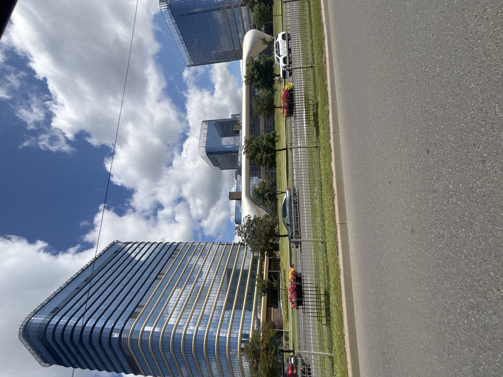 A wide, low-angle outdoor shot captures a modern cityscape with distinctive architecture under a bright, cloudy sky. In the foreground, a grey asphalt road dominates the lower half of the image. Beyond the road, a low metal fence with vertical bars separates it from a strip of green grass.
To the left, a tall, sleek skyscraper with a curved facade made of blue glass and horizontal gold bands rises majestically. Its many windows reflect the sky and clouds. In front of this skyscraper, a dark-colored sedan is visible on the road.
Further into the scene, a long, curved, white architectural element connects two other modern buildings. These buildings are also made of blue glass and have unique, rounded tops. A white SUV is seen driving on the road in front of these buildings.
In the middle ground, between the road and the buildings, are several green trees. Flower planters filled with colorful pink and yellow flowers are placed along the fence. The sky above is a vibrant blue, dotted with fluffy white clouds, some of which are quite large and dramatic. The overall impression is one of contemporary urban development in a well-lit, daytime environment. The architecture strongly resembles the Minsk World development in Minsk, Belarus, particularly the ISMART buildings and the surrounding infrastructure.