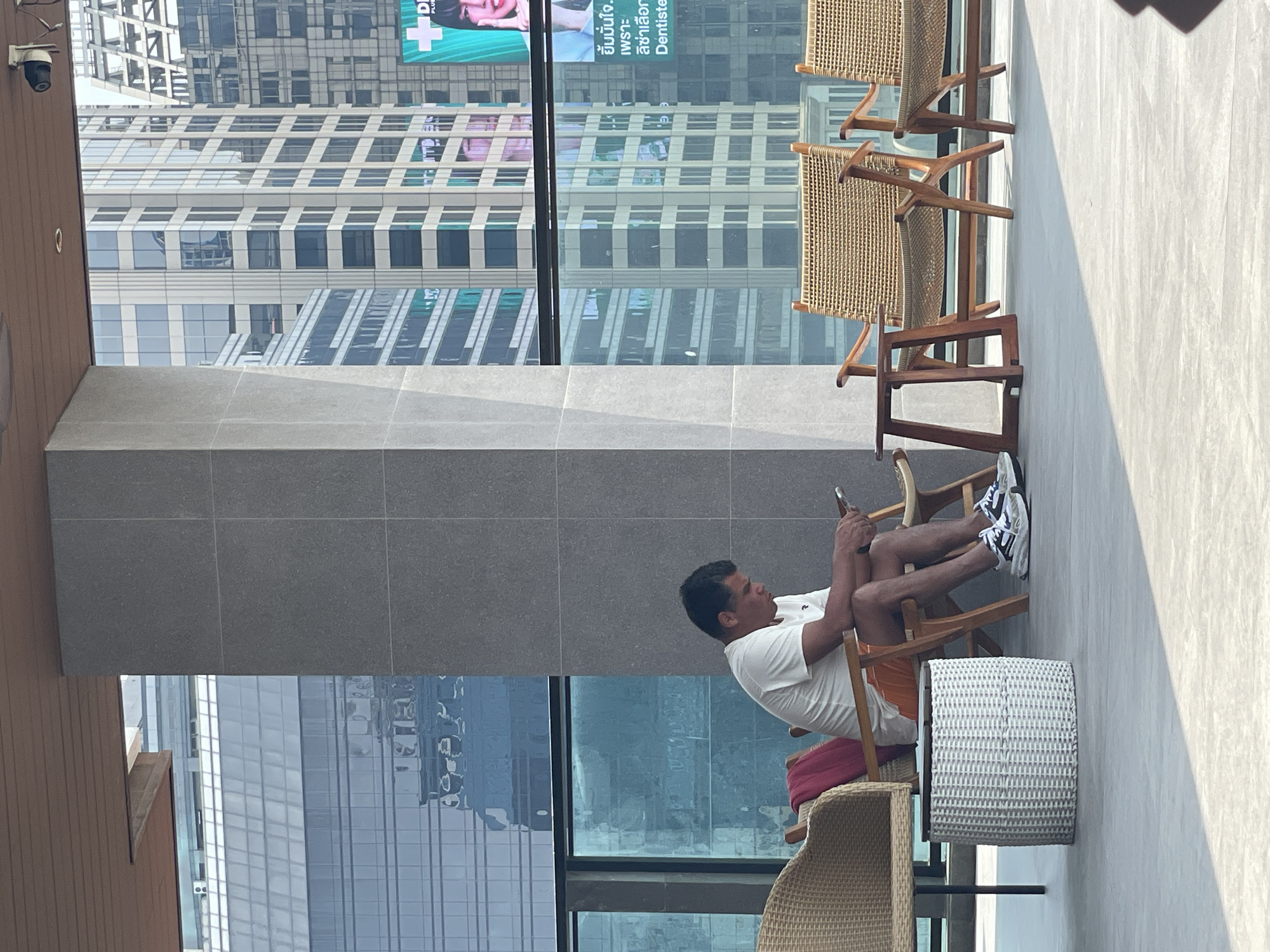 A man sits in a wooden chair, his legs up on another wooden chair, on a balcony overlooking the city of Bangkok, Thailand. The man wears a white t-shirt, orange shorts, and black and white sneakers. He looks at a phone he holds in his hands. Behind him are tall buildings, and to his right is a white wicker-style table. Additional empty chairs are in the background.