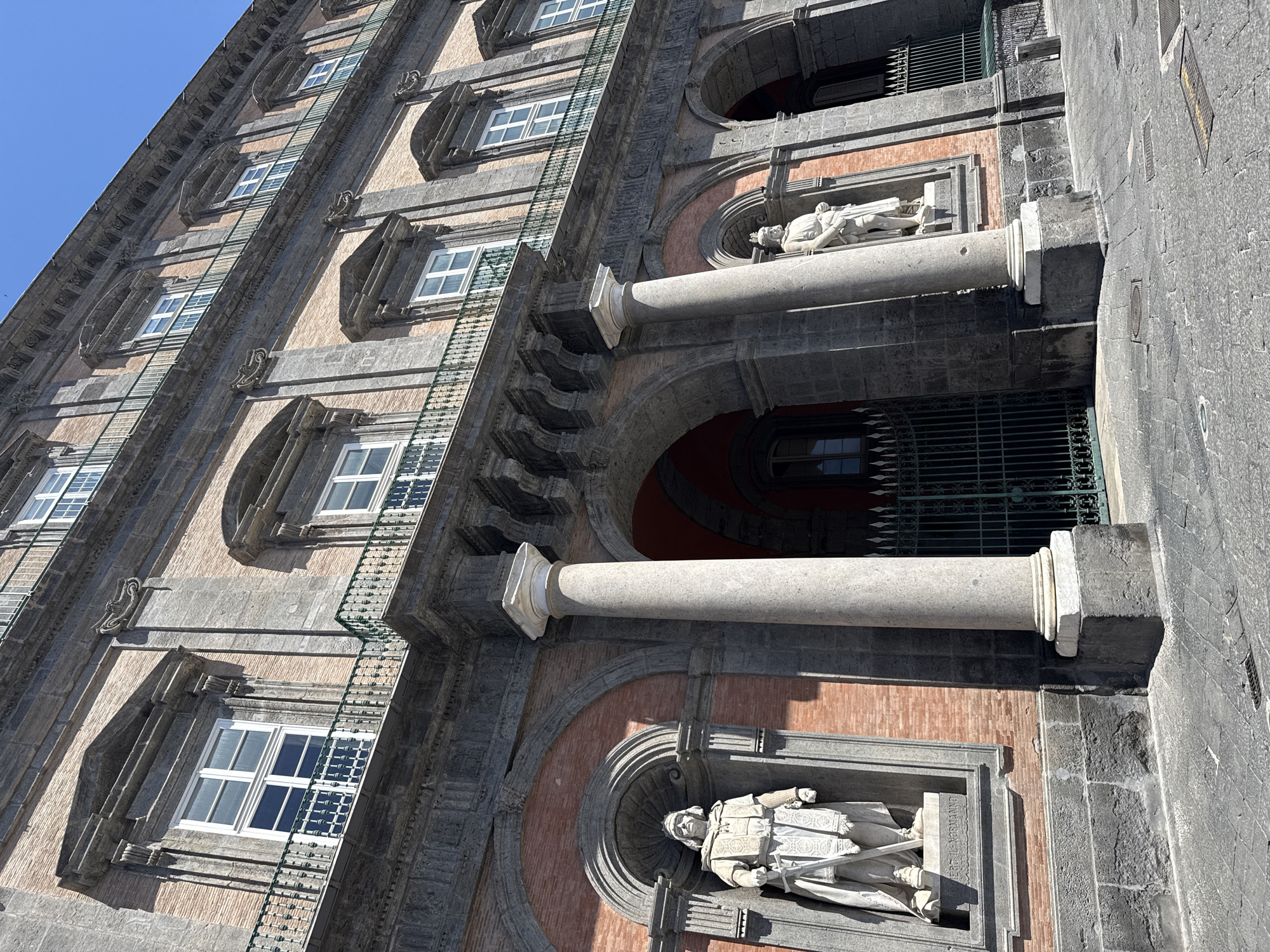 The image shows the Palazzo Reale (Royal Palace) in Naples, Italy, a historic building with a facade made of stone and brick. The palace has multiple stories with arched windows and balconies. Two large stone columns stand in the foreground, flanking an entrance with an ornate green metal gate. Niches on either side of the entrance contain white marble statues of figures in historical attire. The ground in front of the palace is paved with cobblestones. The sky is clear and blue, indicating a sunny day.