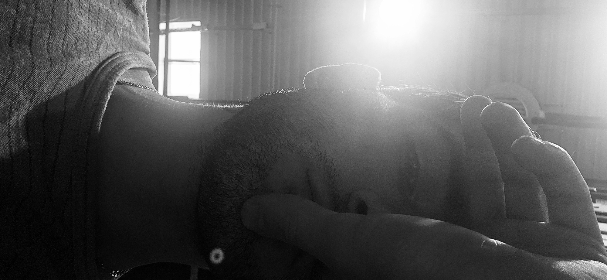 A black and white, horizontal close-up photograph captures a person, likely male, in a contemplative or weary pose. The individual is positioned horizontally, with one hand partially covering their face, possibly shielding their eyes from an intense light source or conveying introspection. Strong backlighting from a bright window or overhead light creates significant lens flare and illuminates the contours of the person's head, face, and shoulder, producing a halo effect around the hair.
The person has short hair or stubble and wears a ribbed or textured garment, possibly a t-shirt or tank top, which is slightly pulled down, revealing part of the shoulder and upper chest. A thin chain necklace is visible on the neck. A small, circular, light-colored object, possibly a piercing or a reflection, is located below the ear on the neck area.
The background features a window with a simple frame, and behind it, corrugated metal walls are discernible. This suggests an indoor, utilitarian, or industrial setting such as a workshop, attic, or storage space. The powerful, direct light indicates a daytime scene, likely with bright weather outside. The high contrast and monochrome palette contribute to a somber, pensive, or introspective mood. The location is Plovdiv, Bulgaria. No visible text is present.