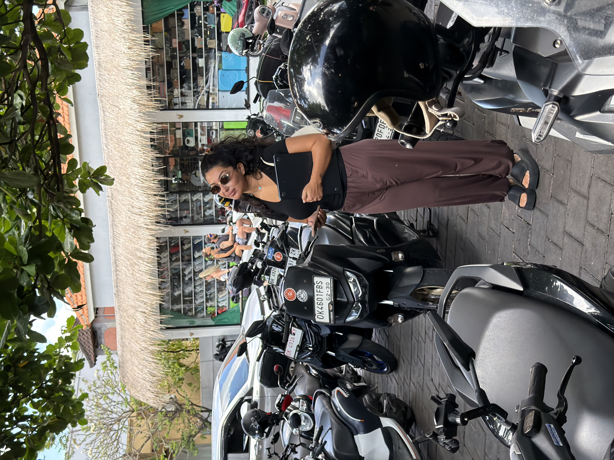 The image captures a vibrant street scene, likely a parking area, bathed in bright daylight. The central subject is a woman standing amidst several parked motorcycles. She has dark, curly hair and wears round sunglasses, a black short-sleeved top, and loose brown pants. She is also wearing black slide sandals and holds a black laptop or tablet in her left arm. Her expression is neutral as she looks slightly to her left.
Numerous motorcycles, predominantly scooters, are densely parked around her. A prominent black motorcycle helmet rests on the handlebars of a bike in the foreground. Several license plates are visible, one clearly displaying "DK 4601 FBS" with "02-30" underneath, and another reading "DK 4916 Y." The "DK" prefix is a common identifier for vehicles registered in Bali, Indonesia.
In the background, a building with a distinctive thatched (straw) roof or awning spans horizontally across the upper middle section of the frame. Behind this, what appears to be a storefront displays various items, possibly bags or other merchandise, visible through its open facade. Lush green foliage from trees frames the left side of the image, with glimpses of a clear sky beyond. The ground is paved with interlocking bricks. The scene conveys a typical busy street environment in a warm climate, consistent with the provided location context of North Kuta, Indonesia.