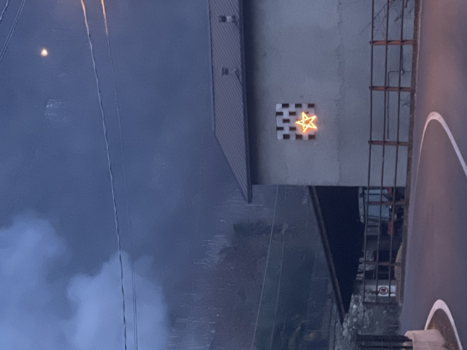 The image displays an outdoor scene in Castione, Italy, captured during twilight or nighttime, characterized by dim, ambient light.
The central focus is a glowing yellow, five-pointed star-shaped light mounted on a light-colored building wall. This star is set against a distinctive checkered pattern of black and white squares. To the left of this wall, part of a building with a dark, corrugated metal roof is visible, extending into the upper left of the frame. Below the wall with the star, a darker area suggests a lower section of a structure or an alleyway, with a metal railing or fence also present on the right.
Further to the right, a paved surface, likely a road or a parking area, is visible with a curved white line painted on it. Overhead, multiple utility lines stretch across the upper portion of the scene.
The sky, which occupies a significant part of the left side of the image, is a muted blue-gray, filled with large cloud formations, particularly dense in the lower left. A bright, circular light source, consistent with the moon, is visible in the upper left sky.
The scene is devoid of people or explicit activity, conveying a quiet and still atmosphere. There is no visible text in the image.