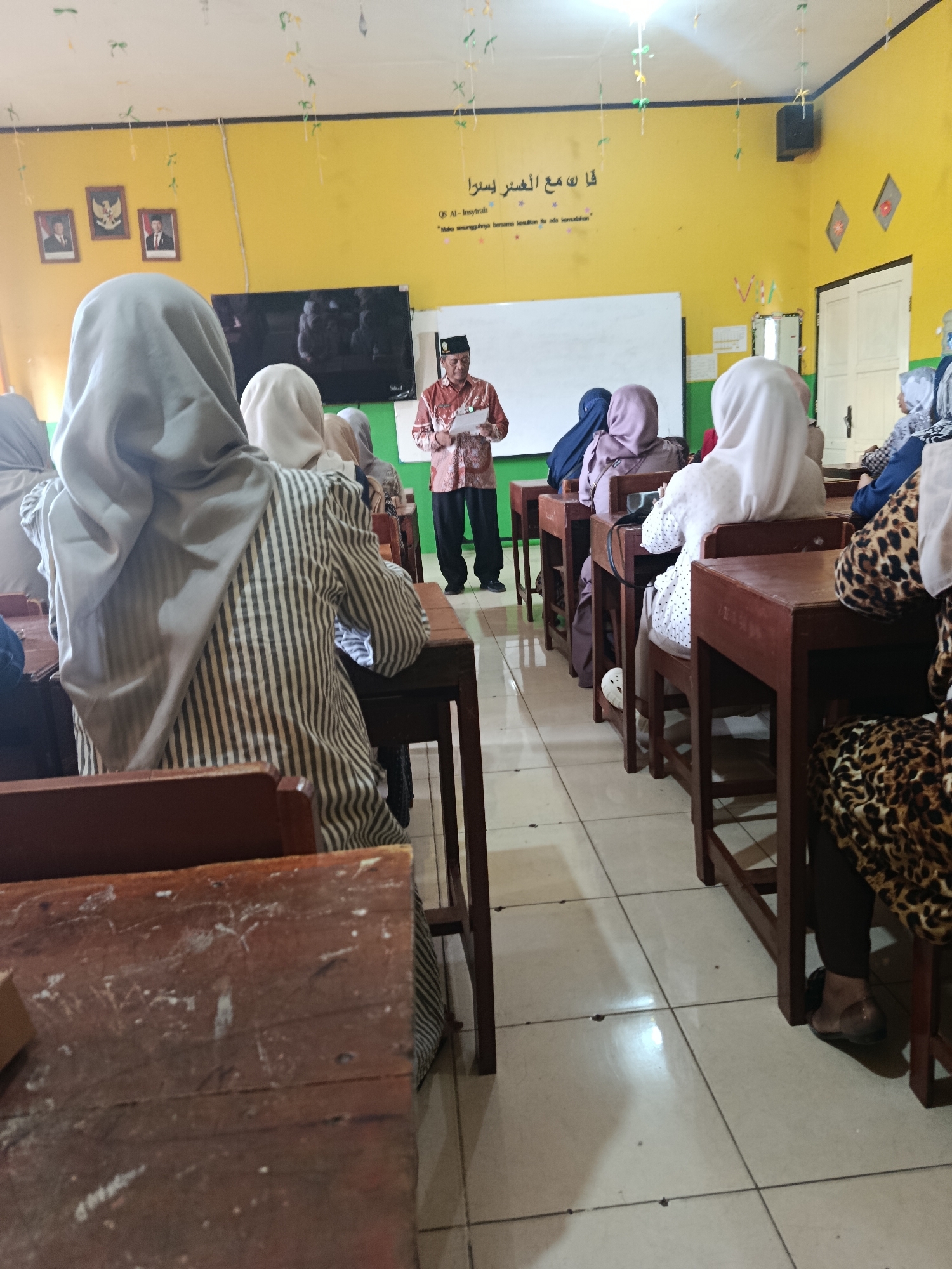 The image depicts a classroom scene in Kawunganten Lor, Indonesia, where a man in traditional attire and a cap is addressing a group of students, mostly young women wearing hijabs. The man is standing at the front of the room, holding a paper. The students are seated at wooden desks, facing the speaker. The classroom has yellow walls with some decorations and Islamic calligraphy. A whiteboard is visible behind the speaker. The overall atmosphere appears to be an educational or religious gathering.