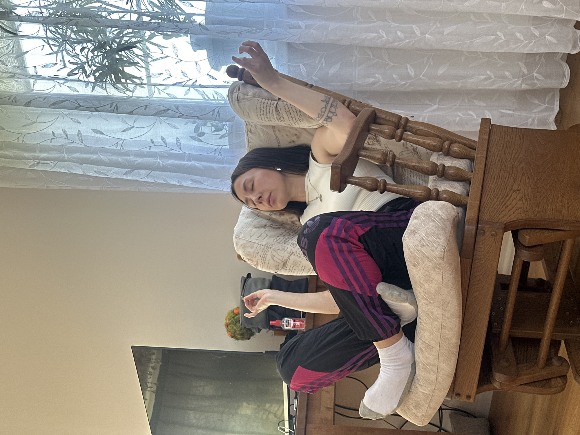 A young woman with dark hair is asleep in a wooden rocking chair. She is wearing a white tank top and black pants with pink stripes. Her left arm is raised above her head. A window with sheer curtains is behind the chair. A television is partially visible in the lower left corner of the frame. A small bottle and a decorative object are on a table to the right of the television. The location is Русский Вожой, Russia.
