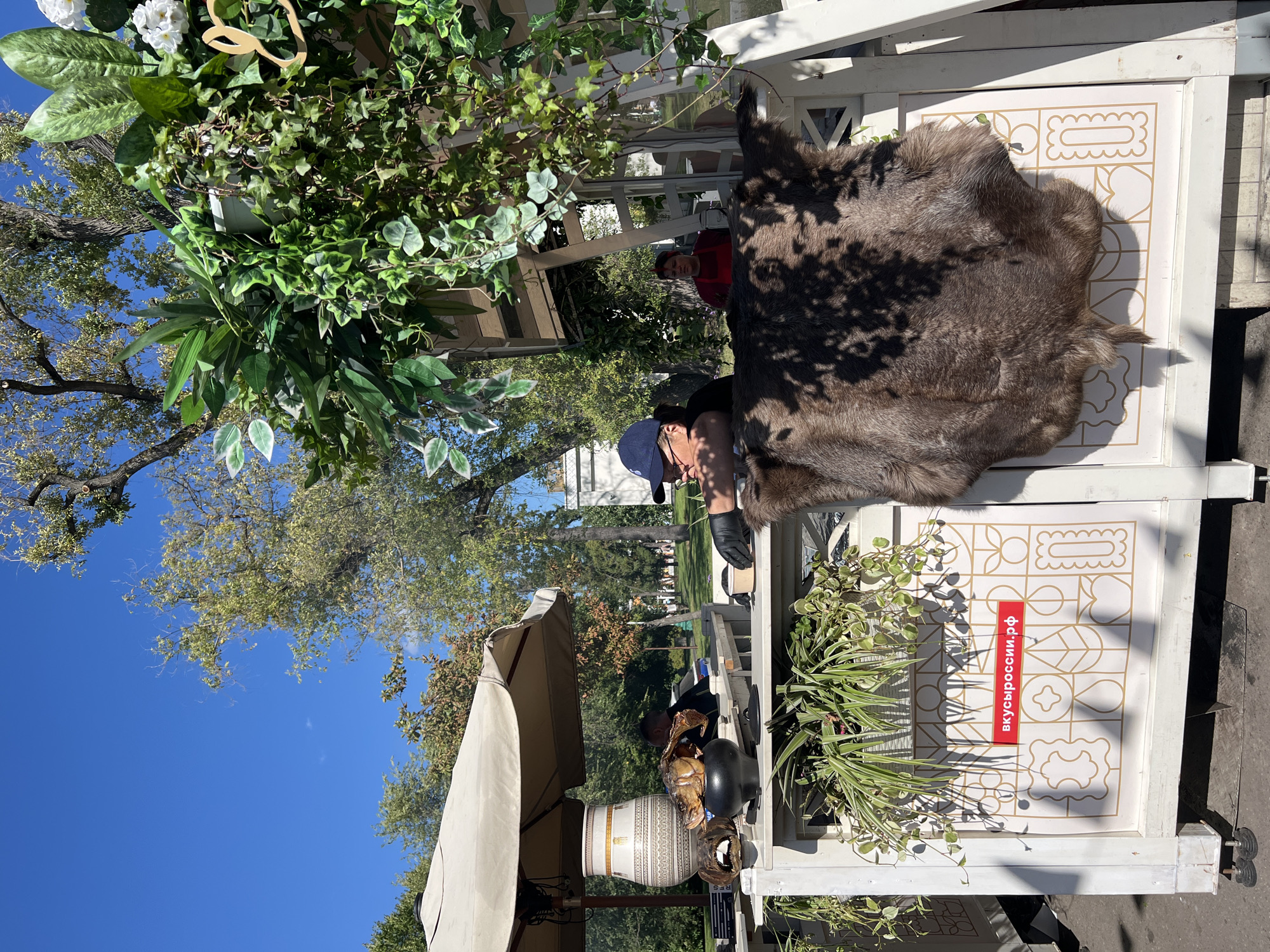 A scene at an outdoor food stall in Moscow, Russia, on a clear day with a bright blue sky. The stall features a white wooden facade adorned with potted plants and a decorative fur rug hanging on the front. On the left, an umbrella shades a table where a large decorative vase and a cooked fish in a pot are displayed. Two individuals are visible behind the counter: one wearing a blue cap and black gloves, and another in a red apron. A red sign with Russian text, "вкусроссии.рф," is affixed to the facade. The background consists of trees and other park-like scenery, suggesting a public outdoor space.