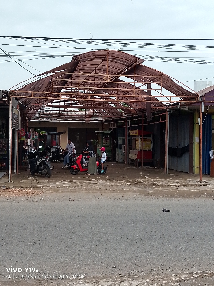 The image shows a partially collapsed metal-framed awning in the village of Haurgeulis, Kecamatan Haurgeulis, Indonesia (latitude: -6.415, longitude: 107.958). Four individuals are visible: two adults and two children. The adults are standing, one is wearing black clothing, and the other is wearing a long, light-colored skirt and a headscarf. Both adults are looking towards the right. The children are both wearing helmets and riding red mopeds, looking straight ahead. Two other mopeds are parked, one with a helmet on the seat. The ground is a mix of dirt and gravel, with one black shoe lying in the middle of the street. The photo was taken on a cloudy day. The awning is partially collapsed with one side of the roof still intact.