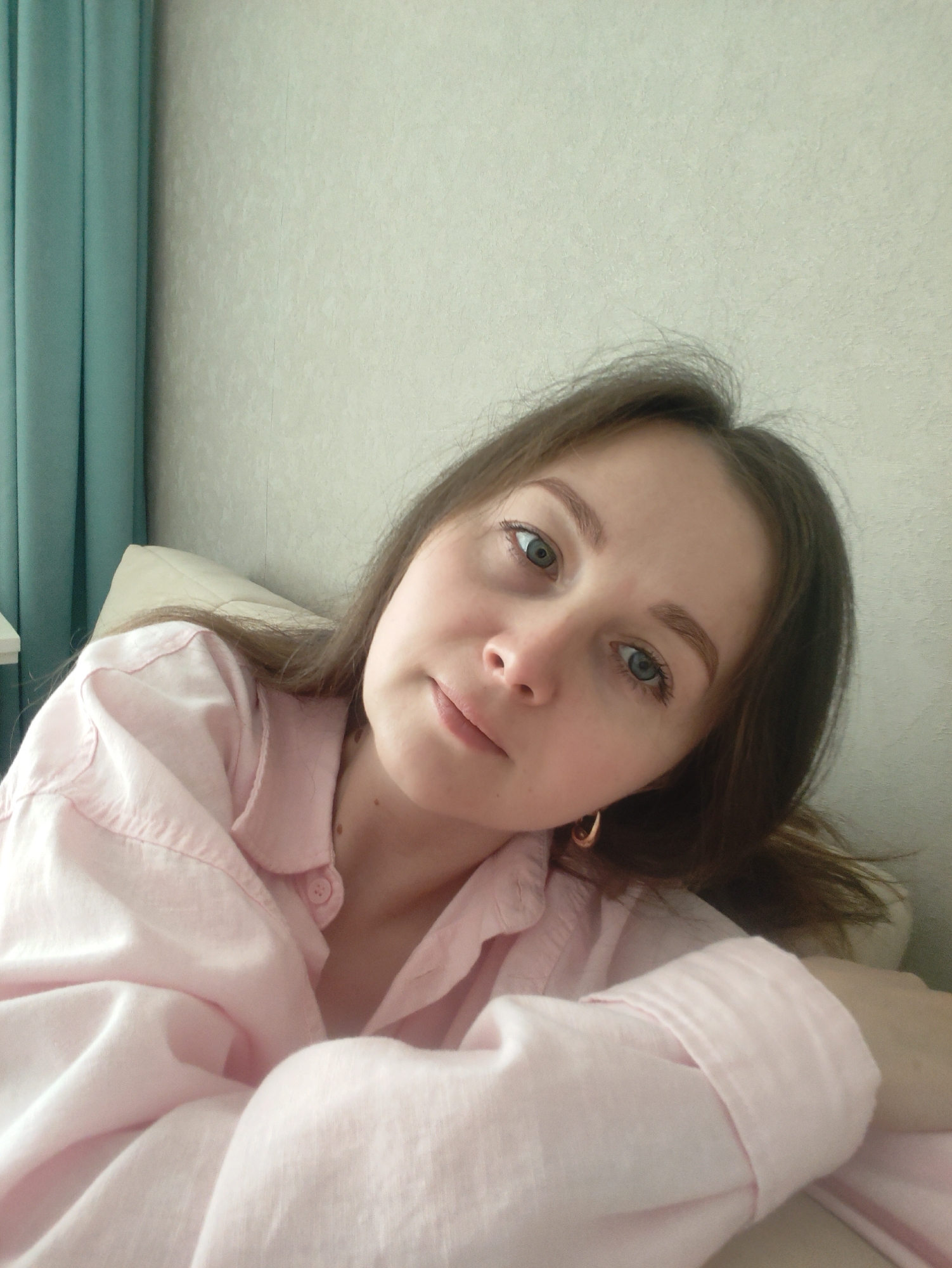 A young woman is captured in a close-up, slightly angled shot, appearing to be taking a selfie or being photographed from an intimate perspective. Her head and upper torso are visible, framed against a simple indoor background. She has long, light brown hair, which falls around her shoulders. Her eyes are a striking blue-green, and she has a calm, neutral, or slightly pensive expression as she looks directly into the camera. There are no prominent blemishes, and her skin appears clear, with a few small moles visible on her neck/shoulder area. She is wearing a light pink, long-sleeved button-up shirt, possibly made of linen or cotton, which could be pajamas or a casual top. The sleeves are rolled up or pushed back, revealing her forearm. A small gold hoop earring is visible on her right ear.
She appears to be reclining or lying down, supported by a light-colored pillow or cushion that is partially visible behind her. To the left side of the frame, a turquoise or light blue curtain hangs, adding a pop of color to the otherwise muted background. The wall behind her is a light, possibly off-white or very pale beige, with a subtle textured pattern. The lighting is soft and ambient, suggesting an indoor setting during daylight hours. No specific activities are ongoing besides posing for the photograph, and no visible text is present in the image. The overall mood is quiet and relaxed.