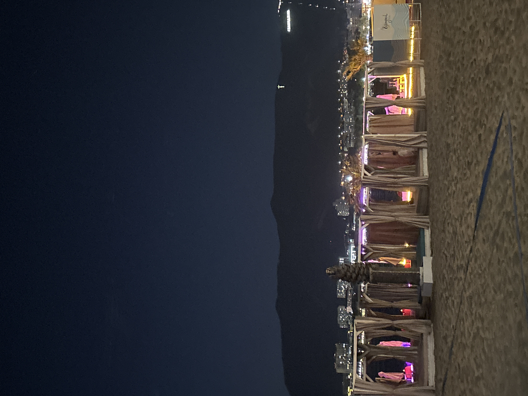 This is a nighttime photograph of a beach with several cabanas set up on the sand. The cabanas are constructed with white wooden frames and draped with light-colored curtains, creating semi-enclosed spaces. Inside some of the cabanas, lounge seating and pink-colored lighting are visible. A single palm tree stands between two of the cabanas. In the background, a city skyline with illuminated buildings stretches across the base of a dark mountain range. A prominent white cross is illuminated on the side of the mountain, and a lighted sign with text that reads "Copacabana" is visible near the right side of the frame. The sky above is a deep, dark blue. The sand in the foreground shows faint blue lines, possibly marking a court for a beach sport.