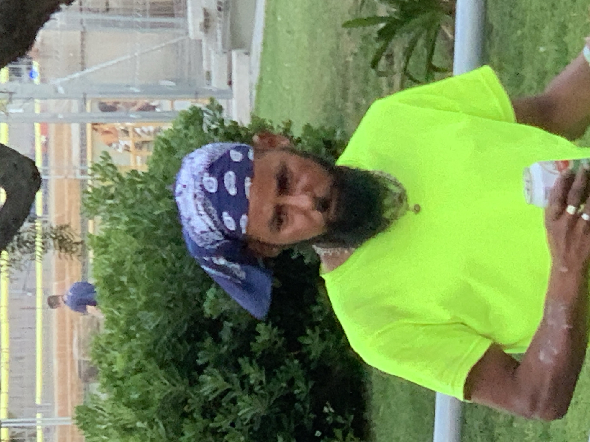 A man with a beard wearing a bright green t-shirt and a blue bandana over his head is holding a can of soda in an outdoor environment. The background shows lush green bushes, a grassy area, and some scaffolding or construction in the distance. It's not possible to determine the location in Barcelona, Spain based on the provided image.