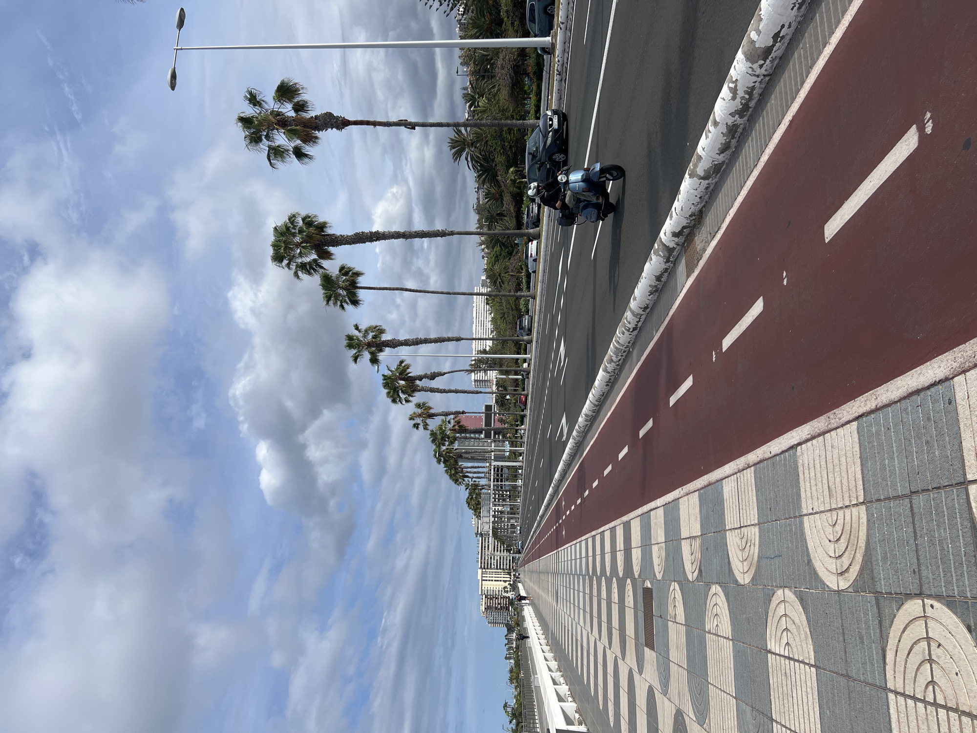 The image shows a coastal scene in Las Palmas, Spain, on a partly cloudy day. The perspective is from the pedestrian walkway. Parallel to the walkway is a red-colored bicycle lane and a gray asphalt road. A person is riding a blue scooter on the road. Several cars are parked next to the road, partially obscured by palm trees lining the street. Buildings are visible in the distance. The walkway is paved with gray and white patterned tiles. Street lights are intermittently spaced along the road.