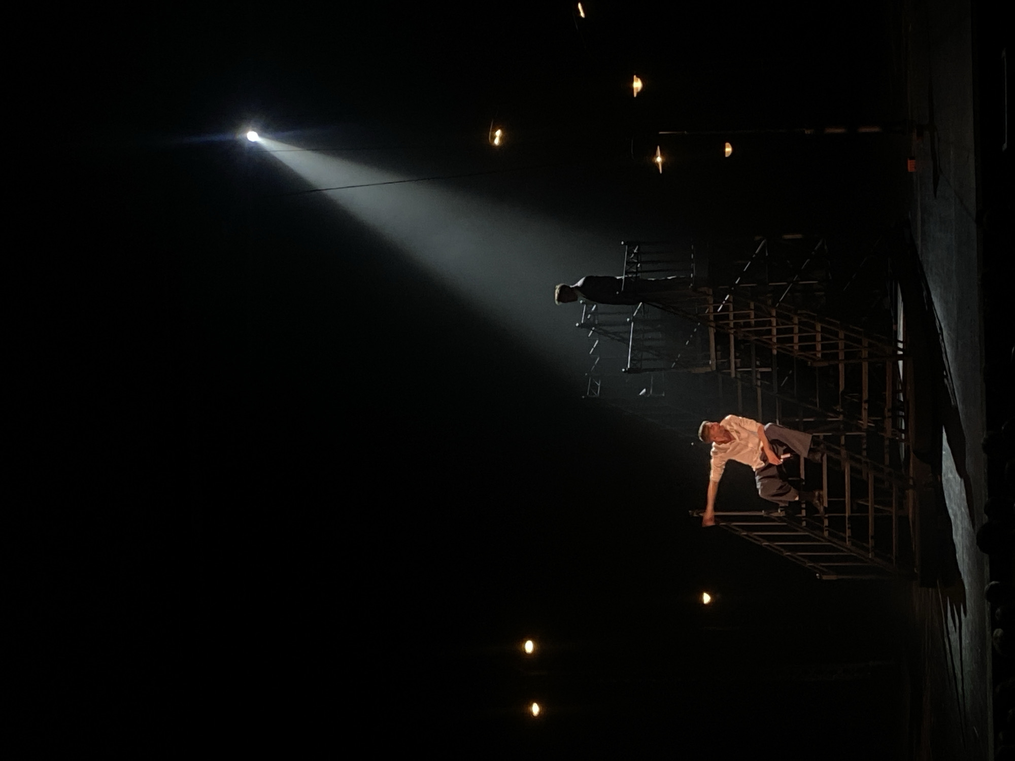 A dimly lit stage features two figures on a multi-tiered metal structure reminiscent of scaffolding or a stairway. A single, bright spotlight illuminates the scene from the upper left. One figure stands atop the structure, partially obscured by shadows. The other figure is lower down, leaning precariously from the structure with an arm extended. The scene is likely a performance taking place indoors. There is no specific information in the image that indicates the location is in the city of Заволжье, Russia.