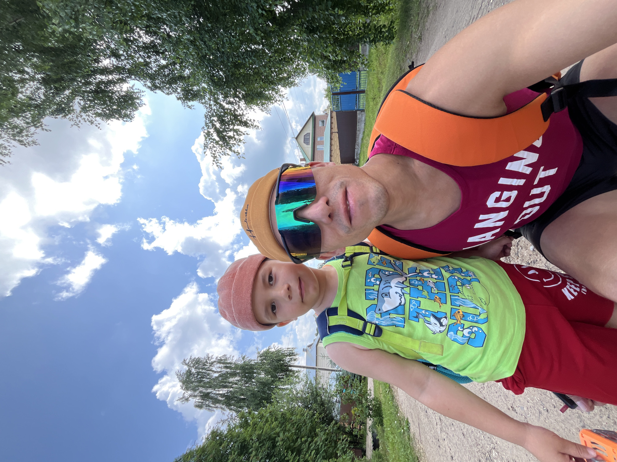 A medium shot captures two individuals, a child and an adult, posing for a selfie outdoors on a bright, sunny day. The child, positioned on the left, wears a light green t-shirt with a shark graphic and red shorts. They have a blue backpack strapped on. The adult, on the right, sports a maroon tank top, black shorts, and an orange backpack. They are also wearing a mustard-colored beanie and reflective sunglasses. The background reveals a dirt road with trees lining both sides. In the distance, a house with a brown fence and a blue gate are visible, suggesting a rural or suburban setting in Chaikovsky, Russia. The sky is a clear blue with scattered white clouds. The sun is bright, casting shadows and creating a luminous atmosphere.