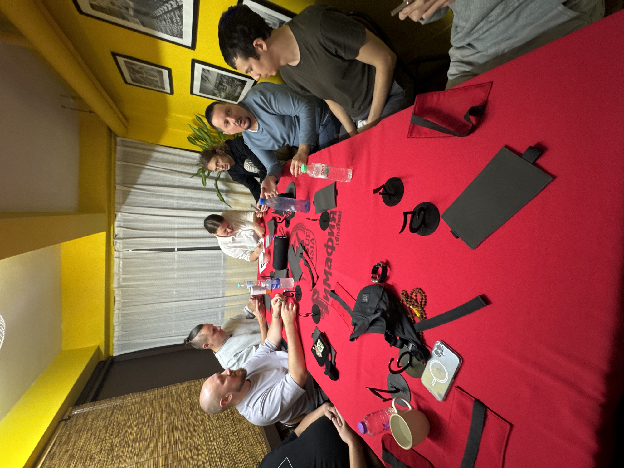 The image shows a group of people gathered around a red table in what appears to be a private room or game space, possibly in Chiang Mai City Municipality, Thailand. The room has yellow walls, with framed black and white photographs hanging on one wall. The table is covered in various objects, including water bottles, what seem to be game cards or accessories, and personal items like a mobile phone and a mug.
Several individuals are visible, seated around the table, engaged in an activity. Their faces show concentration and interaction. One person in a grey t-shirt is looking down at the table, while another in a blue long-sleeved shirt is looking towards the camera. A person in a white shirt is also visible. The presence of what looks like game materials suggests they might be playing a game, possibly a board game or card game, or perhaps engaged in a group discussion or activity.
On the table, there is visible text on some of the objects. One item has prominent red text that appears to be "MAFIA" in a stylized font, along with some Thai script below it. This strongly suggests they are playing the popular party game "Mafia." There are also smaller black circular objects, possibly game pieces, and several red fabric pouches. A beaded necklace is also visible.
The lighting suggests it could be daytime, but the indoor setting makes it difficult to determine the exact time or weather conditions. The overall atmosphere seems casual and focused on the activity at hand. The individuals appear to be adults, and their clothing is casual. The setup with a central table and multiple participants points towards a social gathering centered around a game or shared activity.