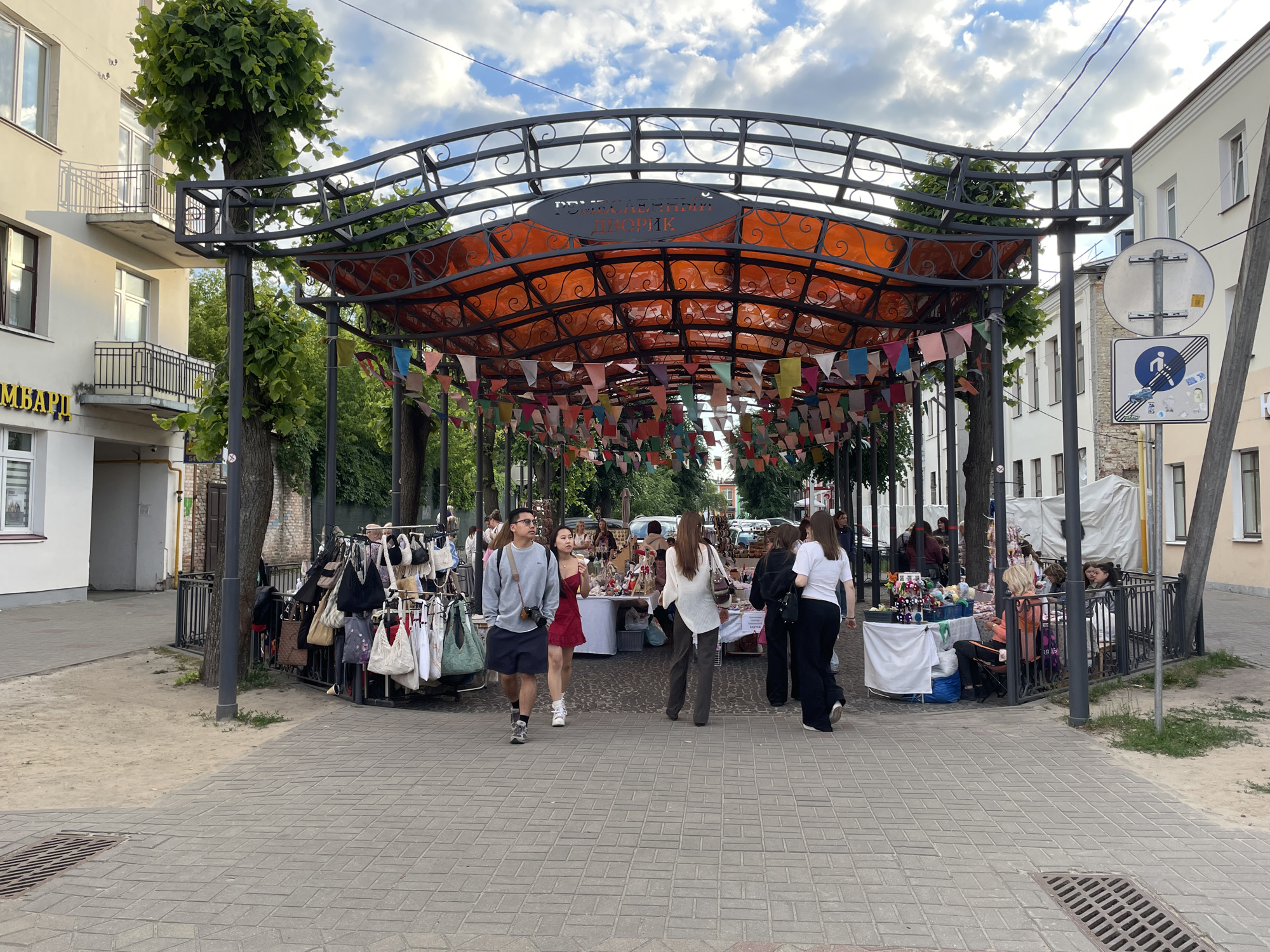 This image depicts an outdoor artisan market, known as "РАМЕСНЫ ДВОРИК" (Ramesny Dvorik), located in the city of Brest, Belarus. The market is situated under a dark metal archway structure featuring a translucent orange roof and numerous colorful triangular pennants suspended beneath it.
The ground consists of light-colored rectangular pavers. Several people are present, either walking through the area or browsing items at various stalls.
In the foreground, a male with short dark hair, glasses, wearing a grey crew-neck sweatshirt and dark shorts, walks from left to right, carrying a camera with a neck strap. Beside him, a female with long dark hair wears a red mini-dress and white sneakers.
Further into the market, a female with long brown hair wears a white long-sleeved top and dark wide-leg trousers, walking towards the stalls. Another female, seen from behind, wears a white t-shirt and dark wide-leg trousers, walking away from the market. Other individuals are visible in the background, including vendors and customers.
Market stalls are set up along the sides, displaying various goods such as bags, knitted items, and small crafts on tables covered with white cloths and on metal racks.
To the left, a multi-story light-colored building is visible with windows and two balconies featuring metal railings. A sign on this building reads "ЛОМБАРД" (Lombard/Pawnshop). Trees with green foliage are integrated into the market structure.
On the right side of the scene, another light-colored building is present. A street pole supports two circular signs: an upper blank sign and a lower sign indicating a "pedestrian zone" or "no vehicles for pedestrians" (a blue circle with a white person walking, bisected by a red diagonal line, also featuring several stickers). Utility wires are visible in the upper right.
The sky above is partially cloudy, with patches of blue. Drain grates are present in the paved ground on both the left and right sides of the foreground, and patches of dirt/sand are visible near the edges of the paved area.