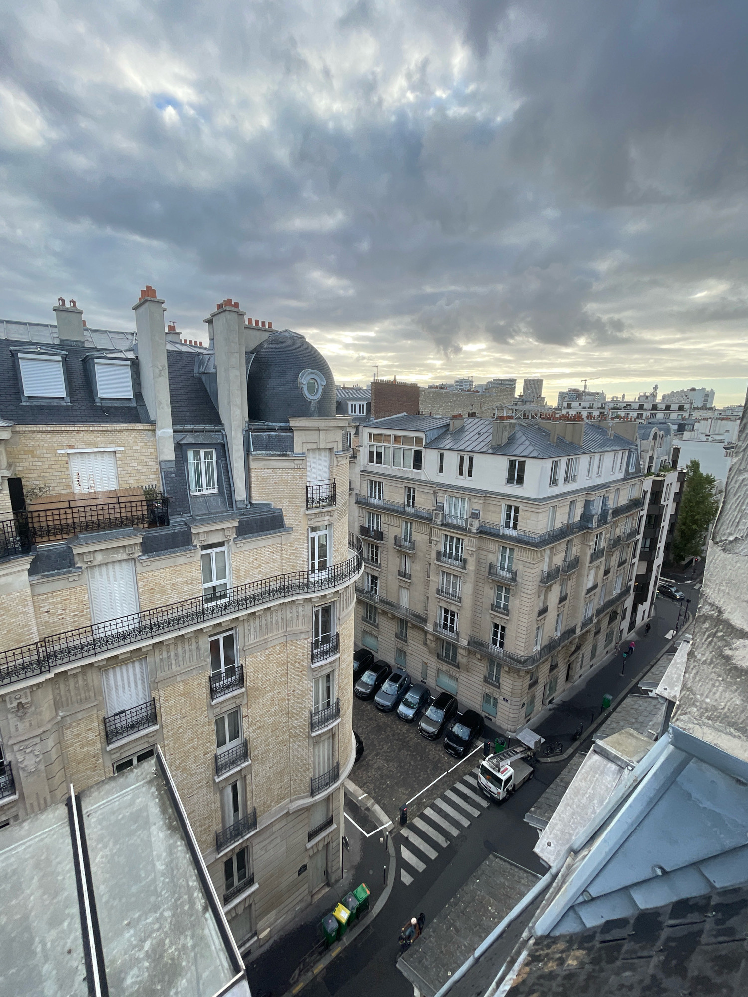 This is an aerial view of a street corner in Paris, France, on a cloudy day. The scene is dominated by Parisian Haussmannian architecture, characterized by cream-colored stone buildings with mansard roofs and ornate balconies. On the left, a curved building features multiple windows with shutters and decorative cornices, topped with several chimneys. To its right, a taller, beige building has a distinct curved facade with more balconies and windows, and a dark, domed roof structure.
In the street below, several cars are parked along the curb, including a row of dark sedans and a white utility truck. A pedestrian crossing is visible, with a few people walking on the street. Three colored garbage bins (green, yellow, and green) are lined up on the sidewalk. The sky is filled with heavy, gray clouds, with some patches of light breaking through. The image captures a typical urban landscape of Paris, with its characteristic buildings and street life under a dramatic sky.