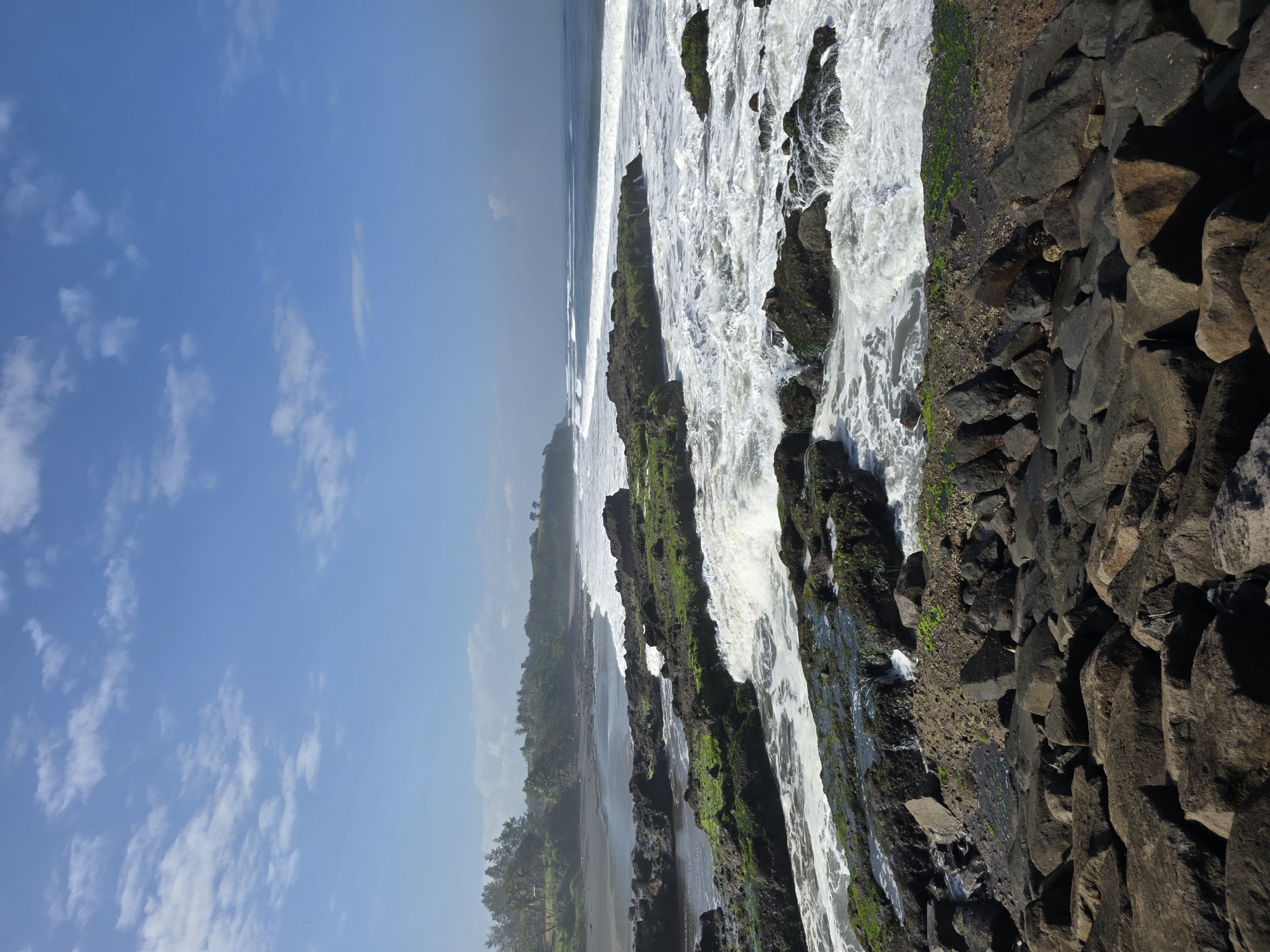 The image depicts a coastal scene with rocky outcroppings and waves crashing against them. In the foreground, large, dark, angular rocks are piled up. Beyond these, a series of dark, moss-covered rocks emerge from the water, with foamy white waves breaking around them and flowing over their surfaces. The middle ground shows a darker beach lined with lush green vegetation and palm trees, with more waves rolling towards the shore. The background consists of a clear blue sky with scattered white clouds, and a hazy horizon where the sea meets the sky. The scene appears to be located in Selemadeg, Indonesia, known for its scenic coastlines. There are no people or animals visible in the image.