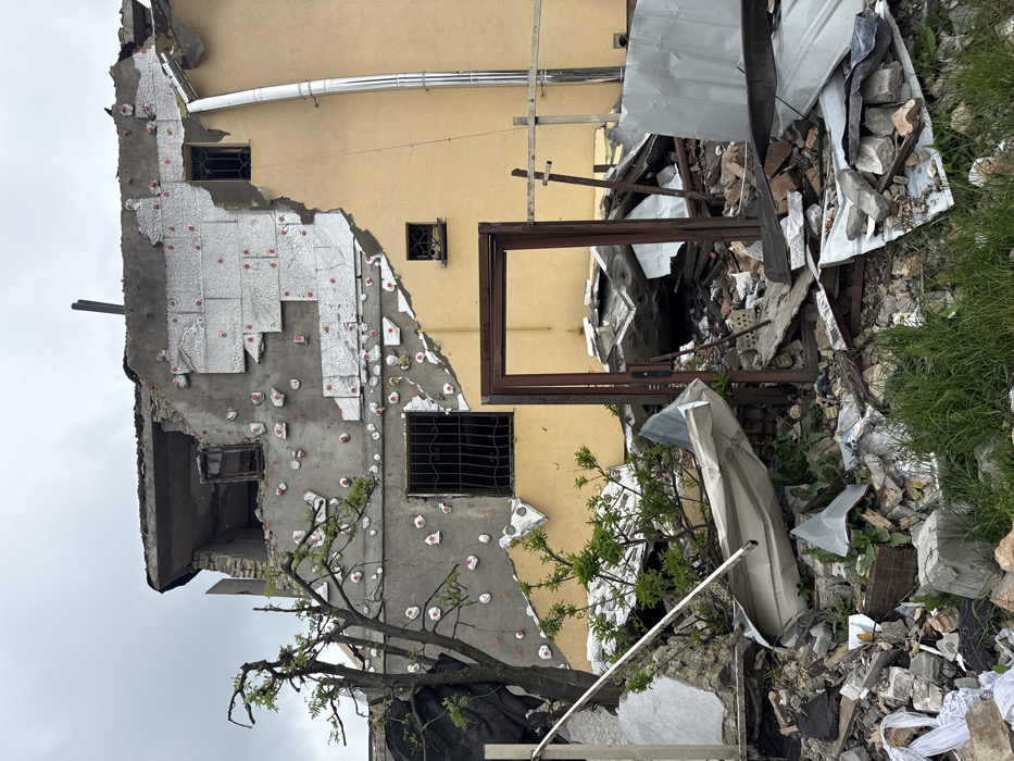 A partially destroyed building stands amidst rubble and debris. The structure reveals layers of construction: an exterior yellow wall and a partially exposed gray wall covered in insulation patches. Windows are present, some with metal bars. An empty, rectangular frame leans against the wreckage. A small tree grows near the base of the building. The scene suggests devastation, possibly from a war or natural disaster.