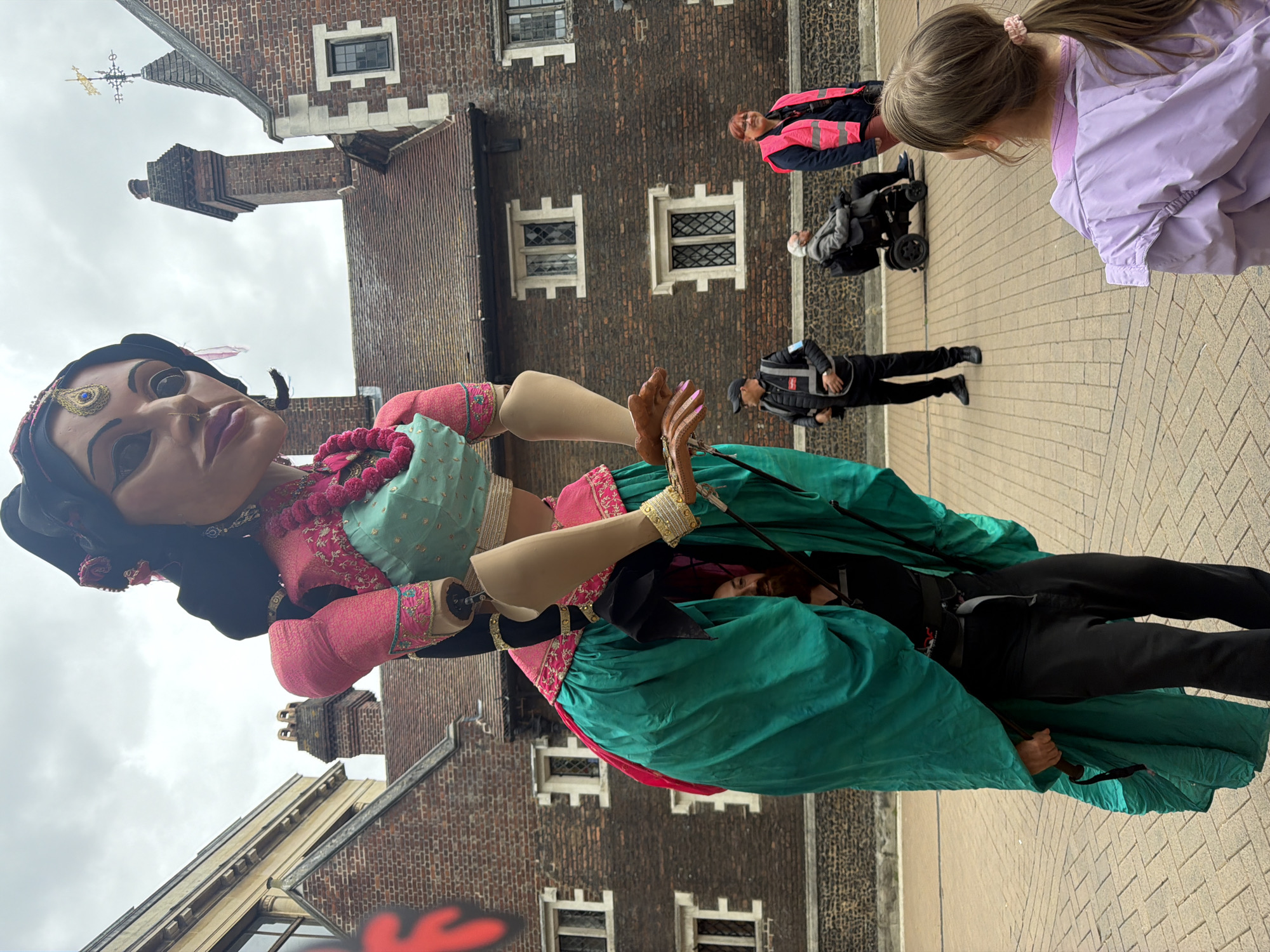 Here is a description of the image:
The image shows a large puppet of a South Asian woman. She is wearing a pink and turquoise top, a long green skirt, and elaborate jewelry. The puppet is operated by a person standing beneath the skirt, and they are walking on a brick-paved area. In the background, there is a brick building with white framed windows and a weather vane. Several people are observing the puppet, including a child with a ponytail wearing a purple jacket, a person in a wheelchair, and other pedestrians. The location appears to be the city of London, United Kingdom, based on the architectural style of the building.