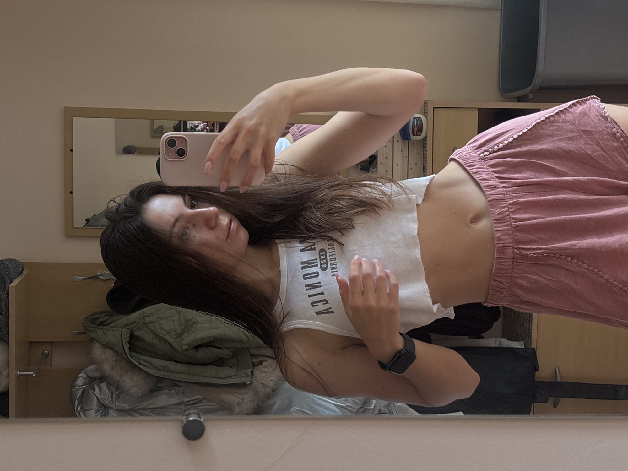 The media file depicts a young woman taking a mirror selfie in what appears to be a bedroom or dressing area. She is positioned in the foreground, leaning back slightly, with her arm extended to hold a smartphone. The woman has long, dark hair and is wearing a white cropped tank top with text that appears to read "SANTA MONICA" in reverse, and pink shorts with an embroidered hem. A black smartwatch is visible on her left wrist.
The background, reflected in the mirror, shows a closet or wardrobe area. A green quilted jacket and a silver puffer jacket are visible, suggesting a cooler climate or season. There are also shelves with various items, including what looks like a bottle of Vaseline and a black bag. The overall lighting suggests it could be daytime, possibly indoors with artificial lighting supplementing natural light. The woman's expression is neutral, and she appears relaxed. The reflection also shows a portion of the room behind her, including a wall and possibly furniture. The scene is set in Varna, Bulgaria, though this context is not visually evident within the image itself.