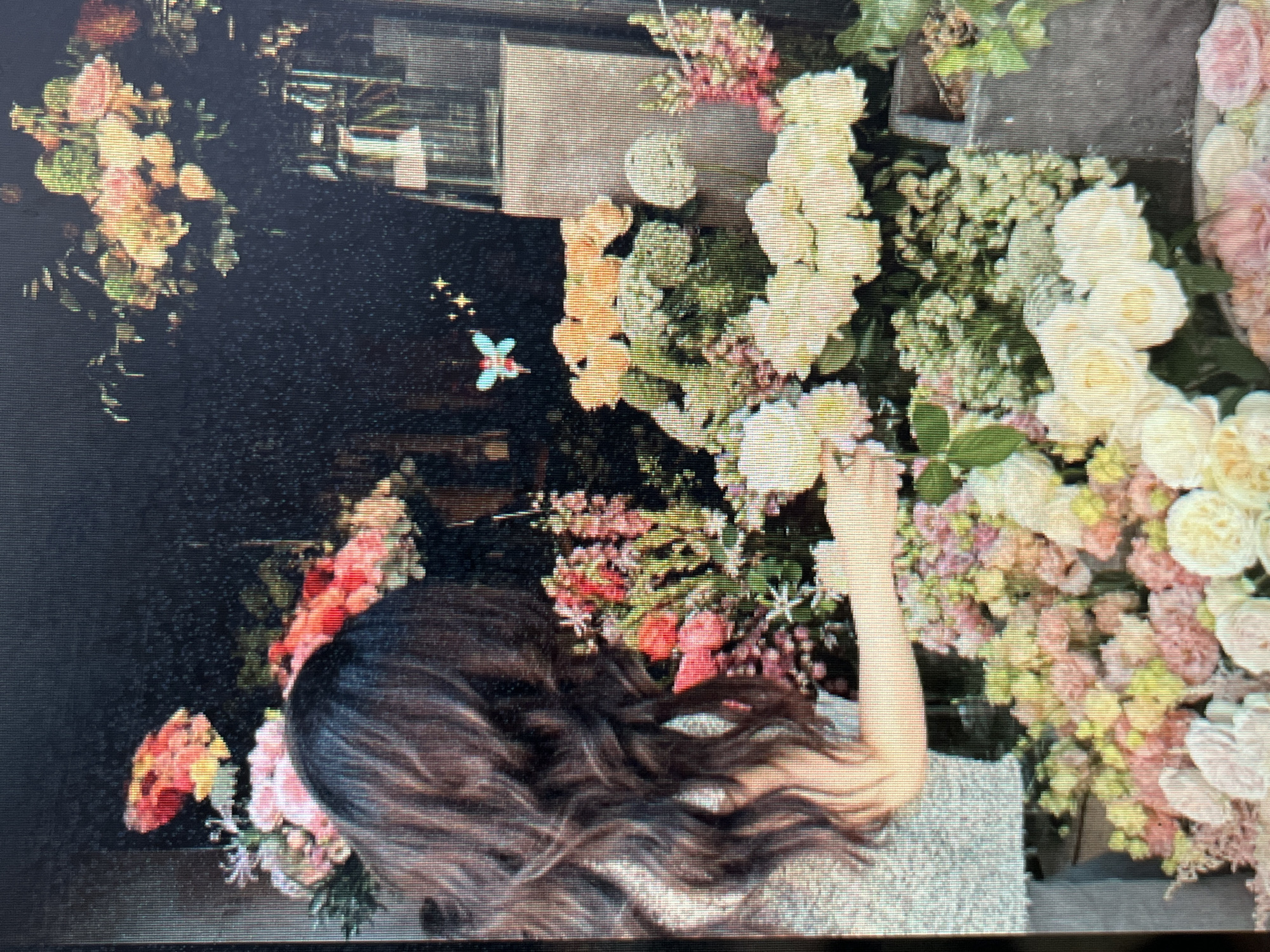 The image depicts a woman with long, dark, wavy hair arranging a large display of flowers. She is wearing a sparkly, light-colored top and is positioned on the left side of the frame, facing away from the camera. The flowers are a vibrant mix of colors including pink, yellow, and white, and appear to be roses and other delicate blooms. Behind her and to her right, more floral arrangements are visible, with some in clear vases. A small, stylized image of a blue and pink butterfly with sparkling accents is floating in the air above the flowers. The background is dark and textured, suggesting it might be a shop or a studio setting. The image is digital and appears to be a photograph or a screenshot. There is no indication of the location in Sejahtera, Indonesia within the image itself.