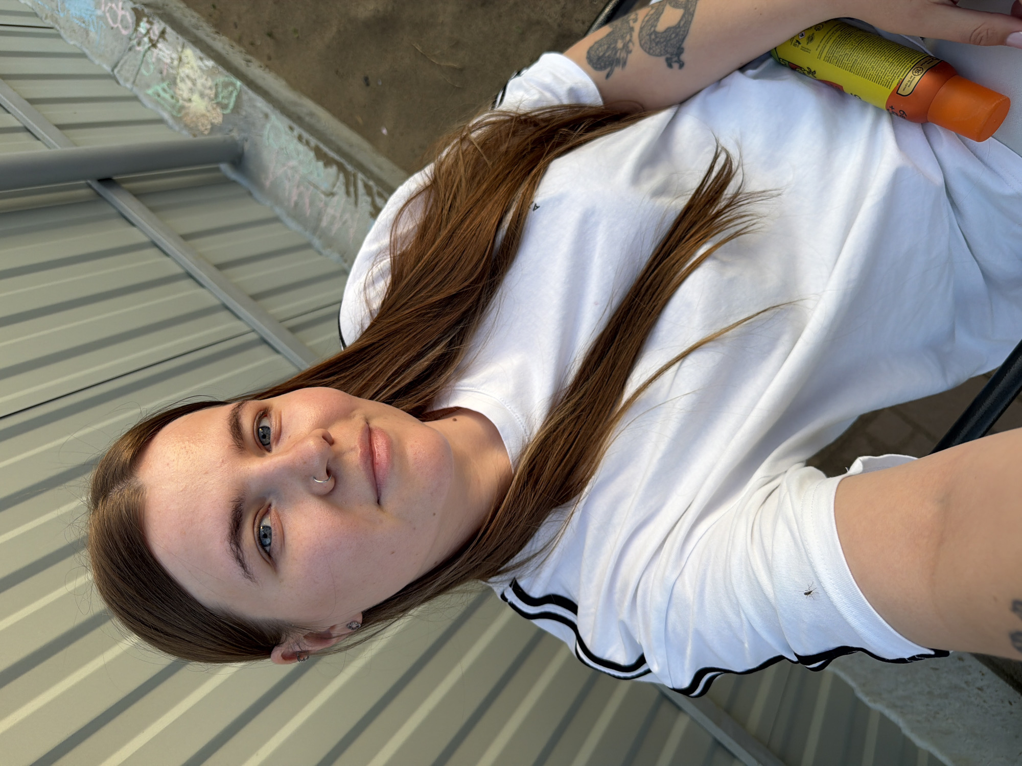 A young woman with fair skin, blue eyes, and light brown hair is photographed from a high angle. She wears a white shirt with black stripes on the sleeves and has a nose ring in her left nostril. Tattoos are visible on her left arm. She is holding a yellow and orange container, possibly a sunscreen or lotion. The background shows a corrugated metal surface, a concrete ledge with graffiti, and metal supports. Given the request for the location, the scene appears to be in an outdoor, urban environment in Chaikovsky, Russia, based on the request's requirements.
