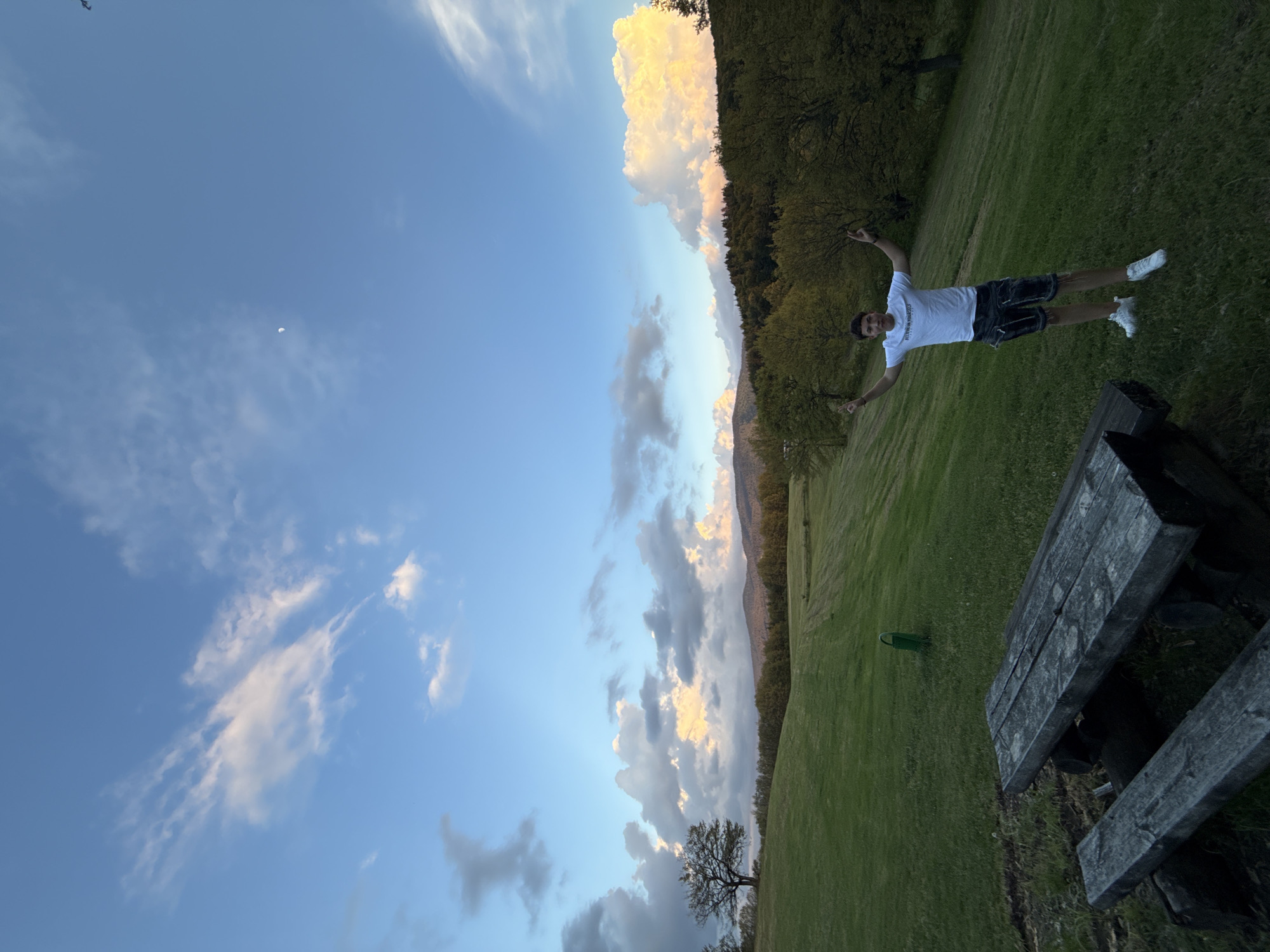 A daytime image captures a man in shorts and a white t-shirt lying on a grassy field, arms outstretched. The field is adjacent to a forest, which transitions into a mountainous landscape. Above, a sky with scattered clouds is visible, with a crescent moon. A wooden picnic table is also in the scene. Based on the location data, this scene is likely in Belchin, Bulgaria.