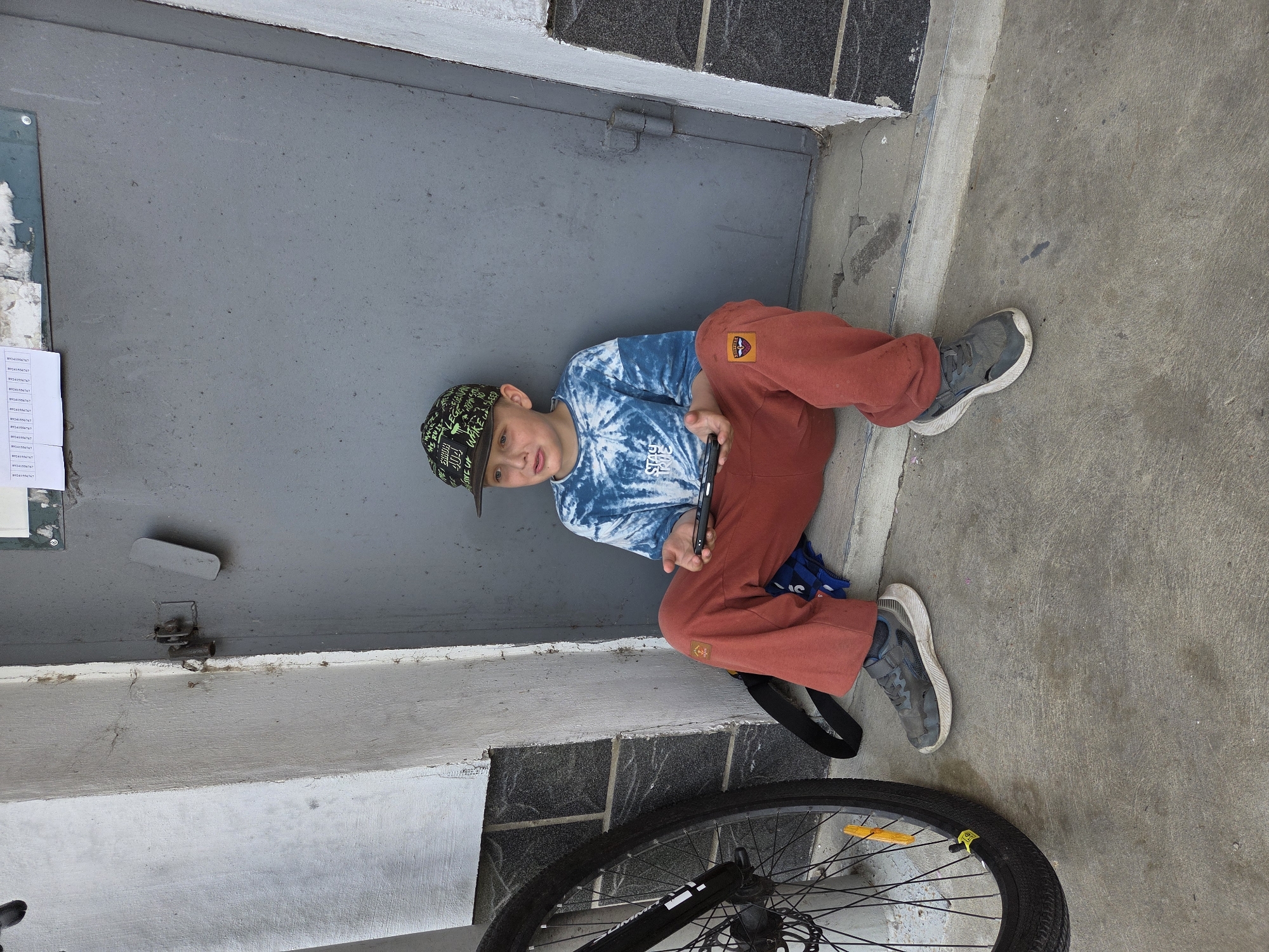 A young boy sits on the concrete ground next to a gray metal door. He wears a patterned cap, a blue tie-dye shirt, and red pants. The boy holds a black object in his hands. Behind him, a black bicycle tire and front forks are partially visible. A backpack is also visible. The location is inferred to be Birobidzhan, Russia, based on the query.