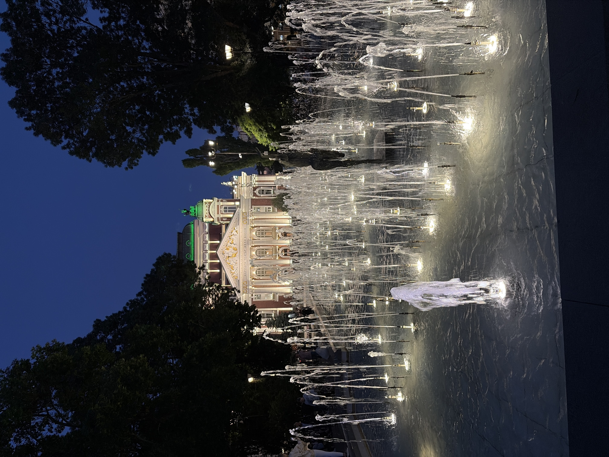 The image depicts a nighttime scene in front of the Ivan Vazov National Theatre in Sofia, Bulgaria. A large, illuminated fountain with numerous jets of water forms the foreground. The water is lit from below, creating a shimmering effect. In the middle ground, people are visible walking around and interacting. Behind the fountain and the people, the grand facade of the national theatre is lit up, showcasing its architecture. Trees line the sides of the scene, and the sky is a deep blue, indicating it is nighttime. Streetlights are also visible, contributing to the illumination of the area. A statue is partially visible amidst the fountain's spray.