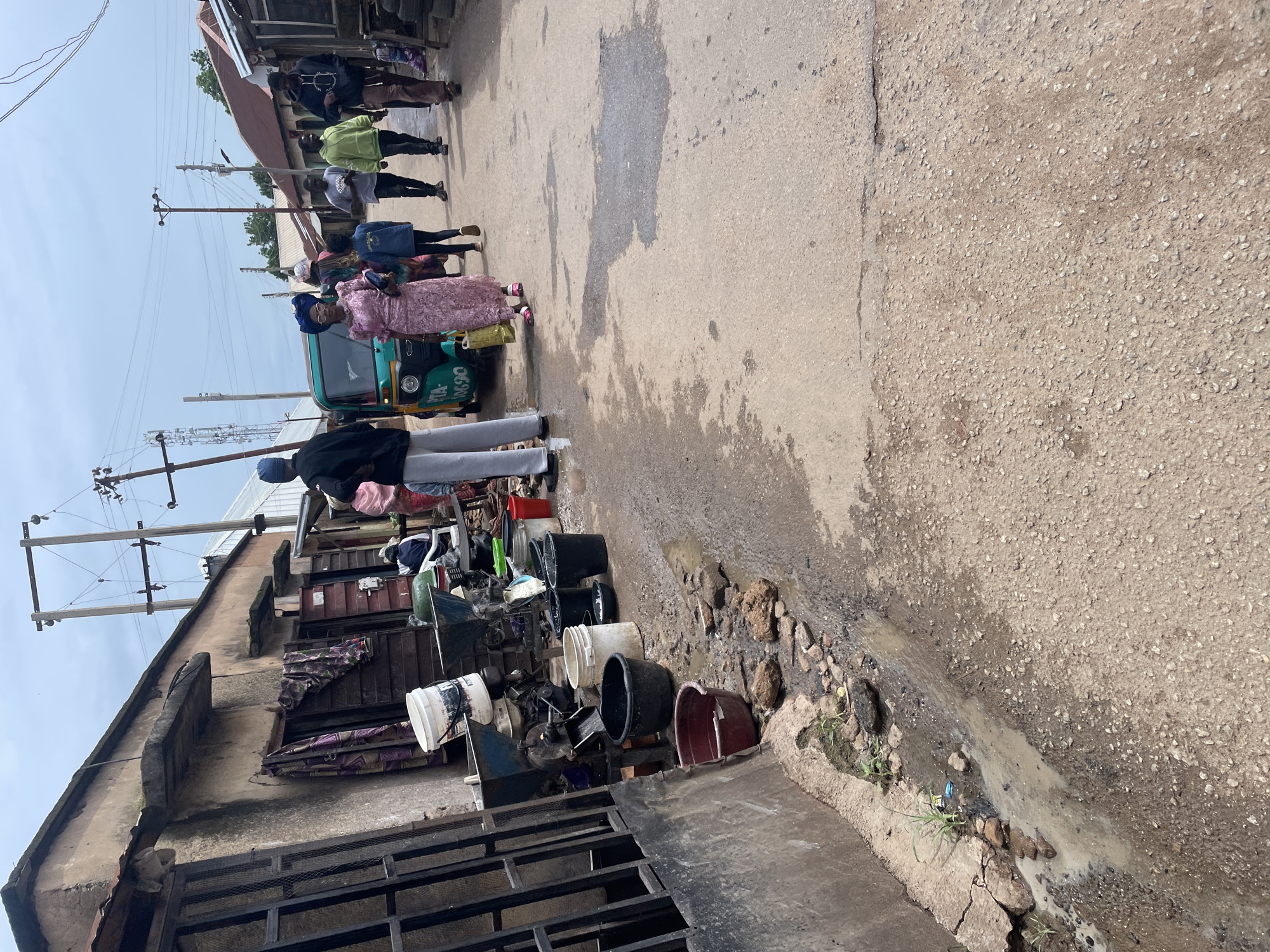 The image depicts a street scene in Jos, Nigeria, likely during daylight hours under an overcast sky. Several individuals are present, including two women standing near a three-wheeled auto-rickshaw, and a group of people further down the street. The woman in the foreground is wearing a pink lace dress and a blue headwrap, and appears to be speaking with the other woman. The auto-rickshaw is green and has the license plate "TA-06690" visible.
The setting is a somewhat dilapidated street with uneven pavement and visible drainage issues, indicated by puddles of murky water and debris. Buildings line one side of the street, with some appearing to be shops or stalls. Various objects are scattered along the side of the street, including numerous buckets of different sizes and colors, some of which appear to be new and some older and weathered. There is also what looks like discarded metal, machinery parts, and other miscellaneous items. Overhead, power lines are visible, supported by utility poles. The overall impression is of a busy, functional, but not affluent, urban environment. The people's expressions are not clearly discernible, but their presence suggests daily life and activity.