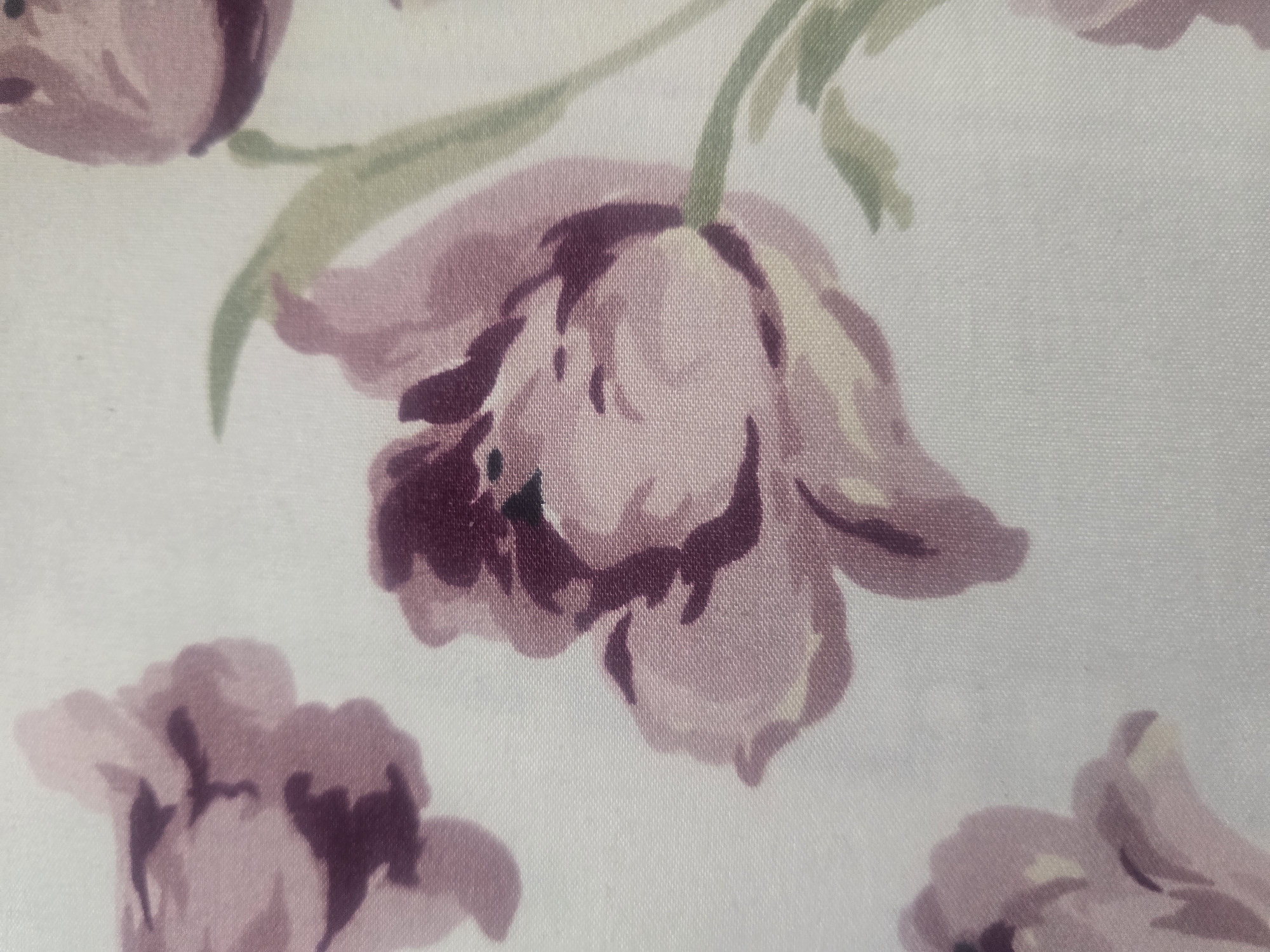 The image is a close-up photograph of a fabric with a floral pattern. The pattern consists of stylized flowers, primarily in shades of pink and deep purple, with some green elements that appear to be leaves or stems. The flowers have soft, painterly edges, suggesting a watercolor or impressionistic style. The background of the fabric is a light, off-white color. The texture of the fabric is visible, showing a fine weave. There are no people or any identifiable elements of a specific city or location within the image. The image focuses entirely on the texture and design of the fabric.