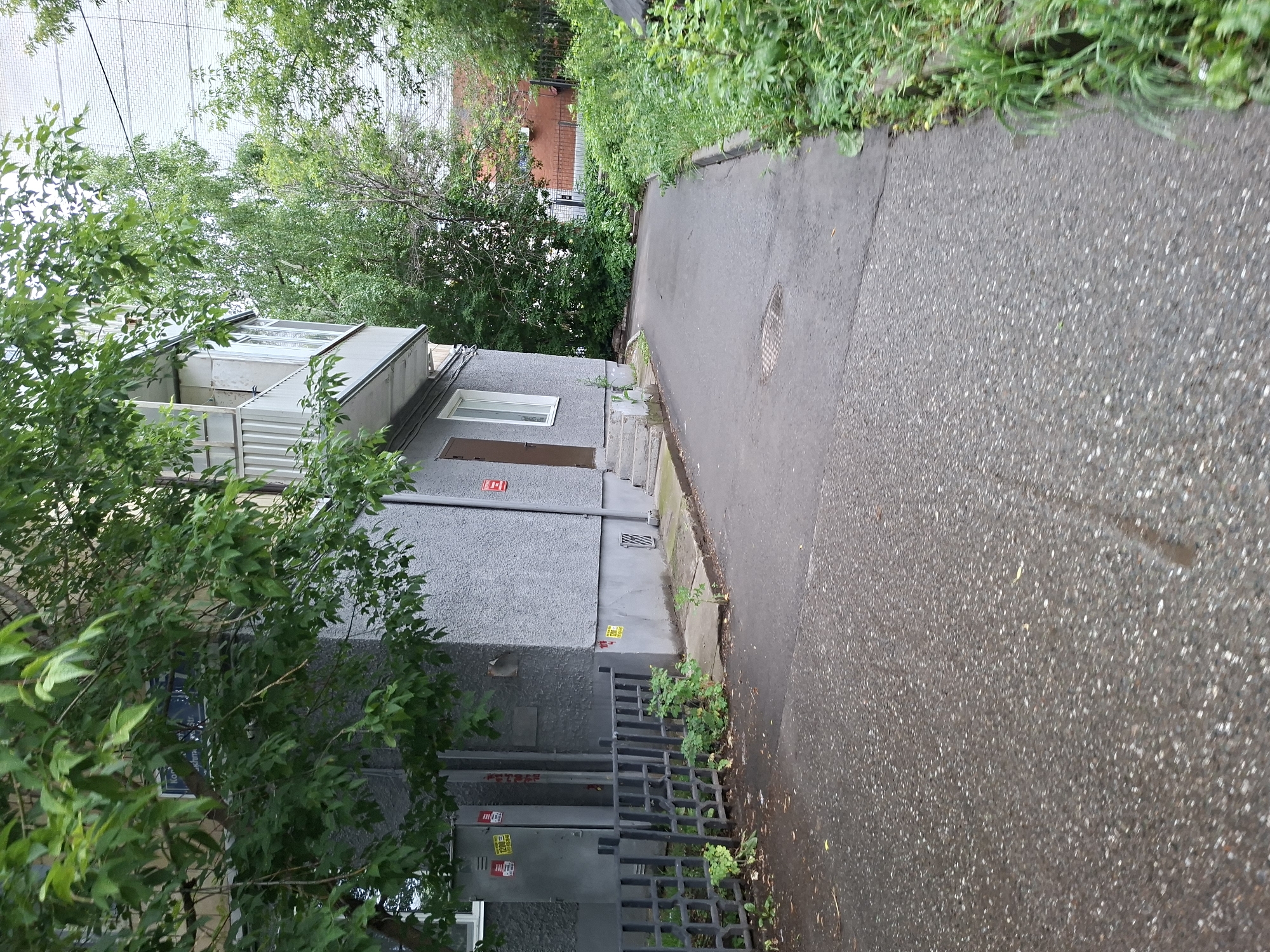 The image shows a view of a residential building and its surroundings in Kazan, Russia. The building is grey with a balcony on the second floor. There is a paved walkway or road in front of the building, with lush green vegetation on the right side. A black metal fence is visible in the foreground. The overall scene appears to be an ordinary urban environment.