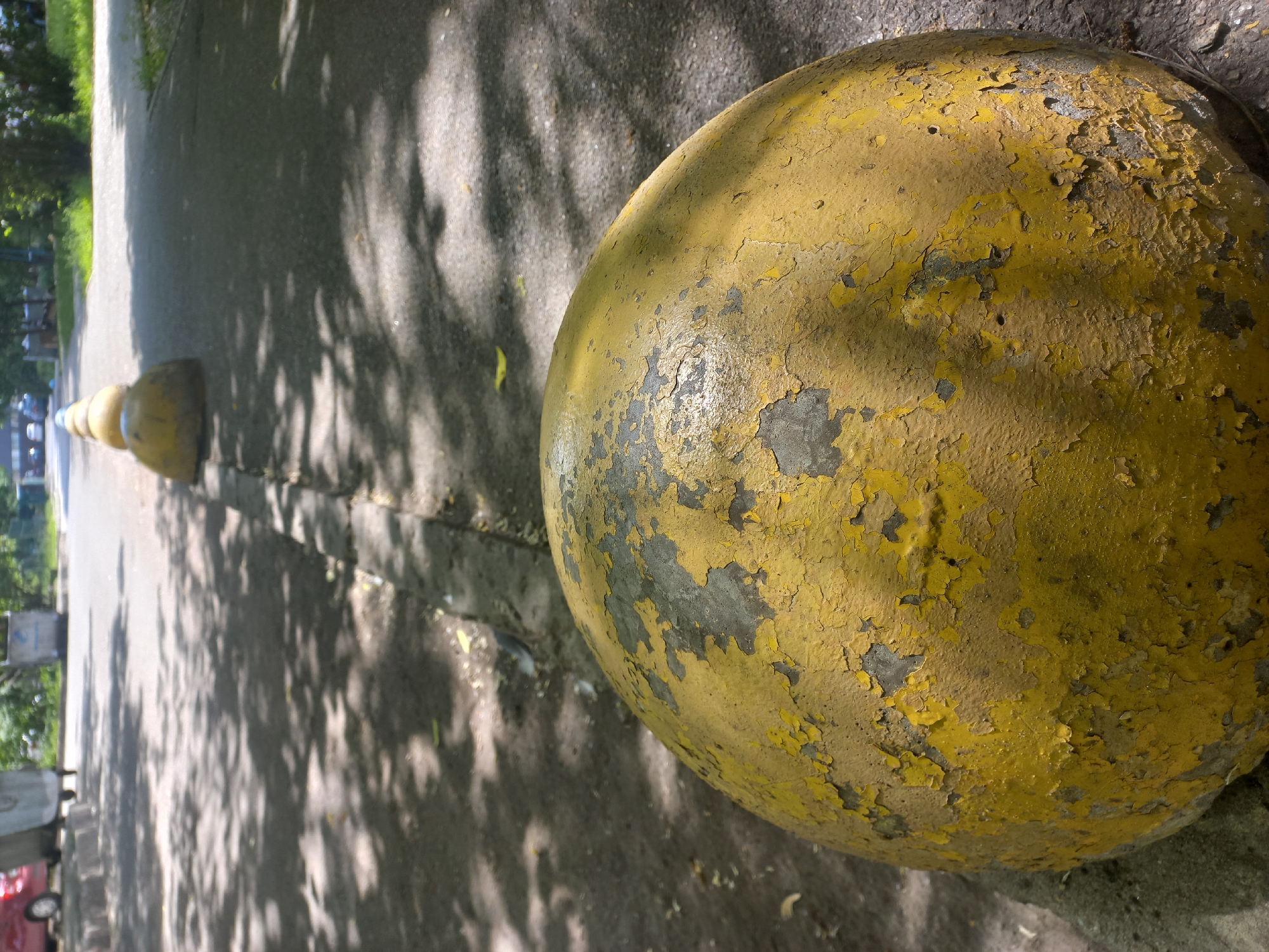 A vertical, eye-level shot captures a row of weathered yellow bollards along a paved path on a sunny day. The dominant subject is a large, spherical yellow bollard in the foreground, occupying the right side of the frame. Its surface shows extensive wear and tear, with large patches of yellow paint peeling off to reveal a darker, possibly gray or black, material underneath, creating a textured, aged appearance.
To the left of this prominent bollard, a line of smaller, similarly yellow, conical bollards recedes into the background, marking the edge of the paved surface. The path itself is dappled with distinct, intricate shadows cast by overhead trees, indicating bright sunlight. Green foliage and trees are visible on the left side of the path in the mid-ground.
In the blurred distance on the far left, indistinct shapes suggest buildings, a red vehicle, and possibly other urban elements, hinting at an outdoor setting like a park, pedestrian area, or roadside. There are no clearly visible people or discernible activity. No legible text is present in the image. The focus is primarily on the texture and condition of the foreground bollard and the interplay of light and shadow on the path.