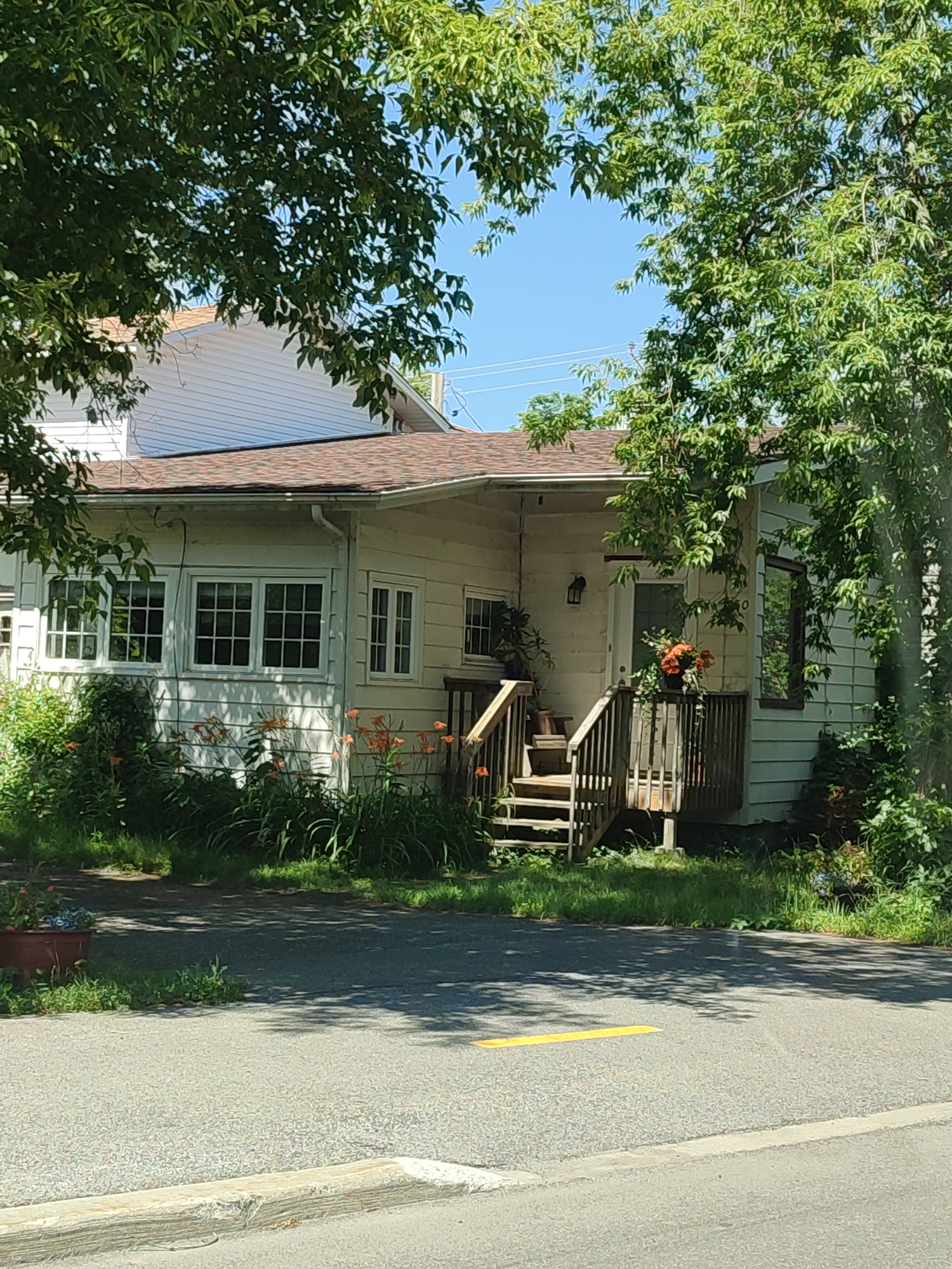 The image shows a single-story, light-colored house with a brown roof, partially obscured by lush green trees. The house has several windows with white frames and grid patterns, and a wooden porch with stairs leading up to the front door. Orange flowers are visible in the foreground, along with other greenery. A portion of another house, with white siding, is visible behind and to the left of the main house. The scene appears to be in Kahnawake, Canada. The foreground shows a paved road with a yellow line painted on it, and a concrete curb. There are no people or animals visible in the image. The environment is sunny, with shadows cast by the trees on the road and the house.