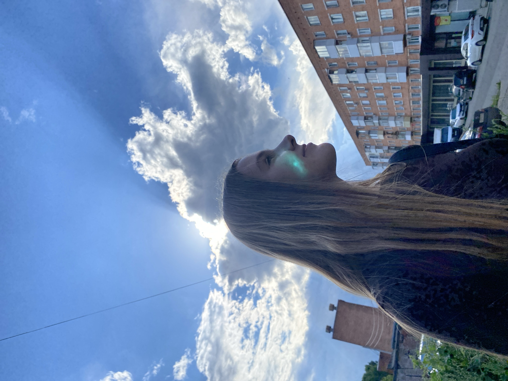 A young woman with long, blonde hair stands in profile, facing right, looking up at the sky. She is wearing a dark, textured top. A green light reflects on her cheek. In the background, a multi-story brick apartment building is visible, with several cars parked on the street in front of it. The sky is blue with dramatic, sun-drenched clouds. To the left of the building, a brick structure with two small towers protrudes. This scene is located in Votkinsk, Russia.