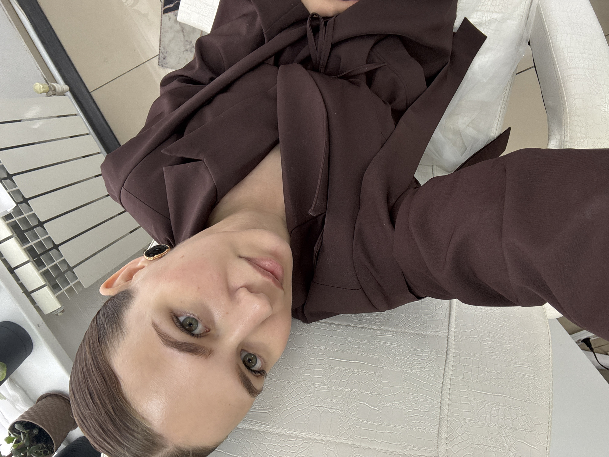 The image shows a close-up, high-angle selfie of a young woman with fair skin and light brown hair pulled back. She is wearing a dark brown blazer over white pants and has a large black and gold earring in her left ear. Her eyes are green, and she has lightly applied makeup. The background suggests an indoor setting, possibly an office or a home, with a white textured chair or sofa in the foreground and a radiator and window with a potted plant visible in the distance. The flooring appears to be tiled. There is no indication of the specific location within the city of Chaikovsky, Russia.