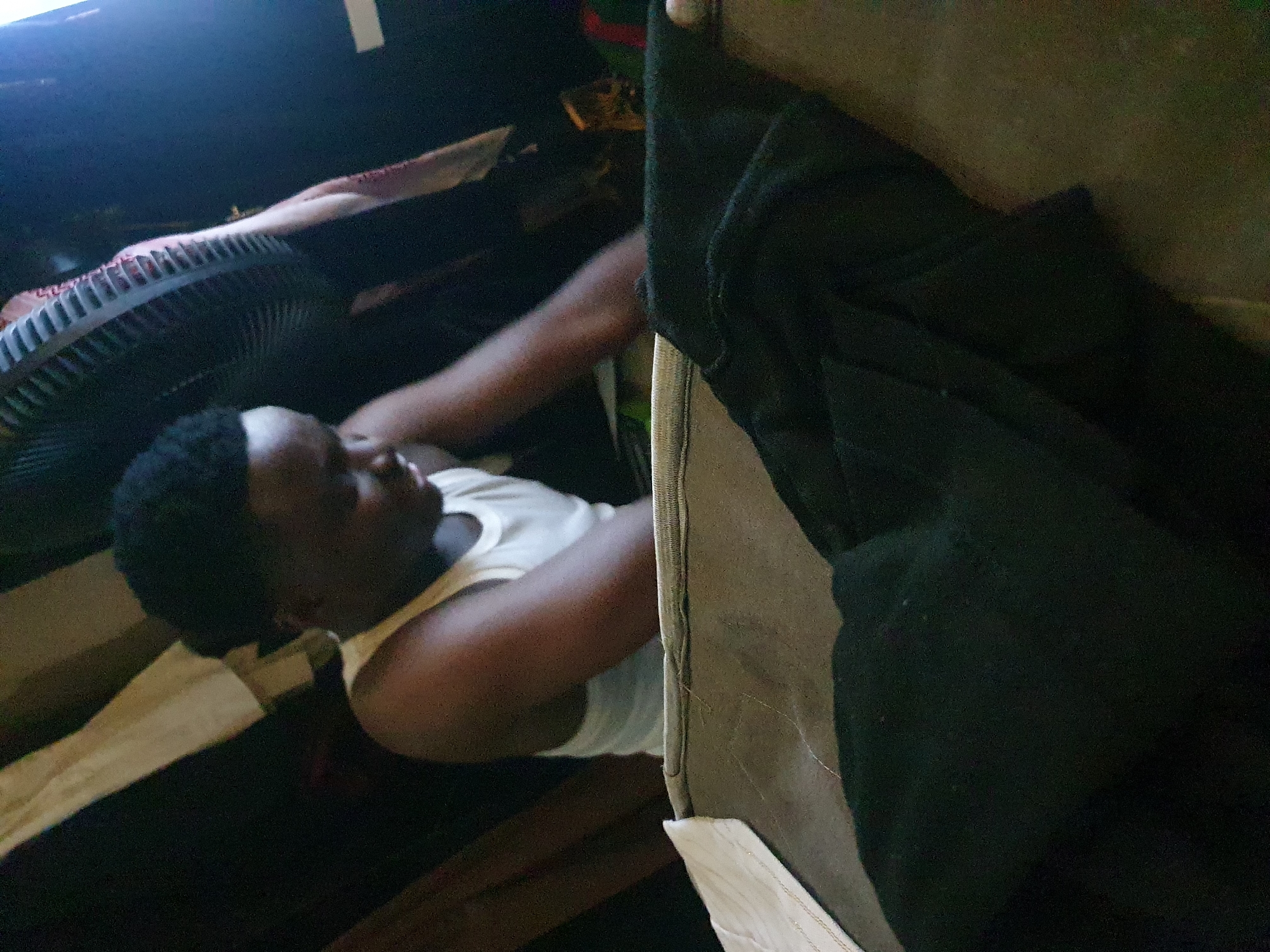 A man lies on a bed, with a fan to his left. The location is a room, presumed to be in Lokoja, Nigeria. The immediate environment includes bed linen, and a dark garment is partially obscuring the view.
