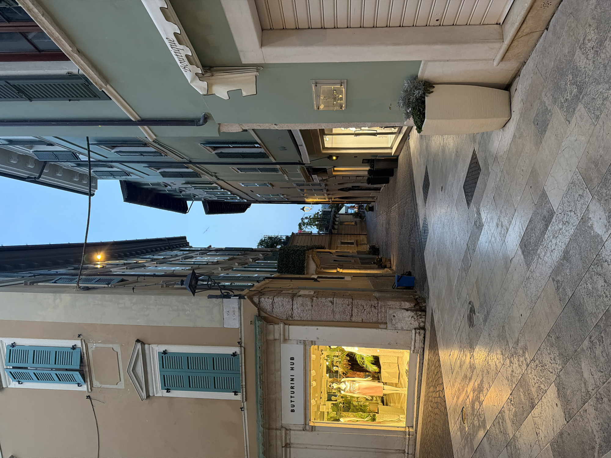 The image shows a narrow street in the historic center of Salò, Italy, at dusk. The street is paved with light-colored stone tiles, and the buildings on either side are tall and narrow, with traditional Italian architecture. The building on the left is painted a light beige and features a shop with a brightly lit window display. The shop's name, "BUTTURINI HUB," is visible above the window. The building on the right is painted a light green and also has a shop with a well-lit interior. The street is empty of people. The sky is a clear blue, and the streetlights are on, casting a warm glow on the pavement.